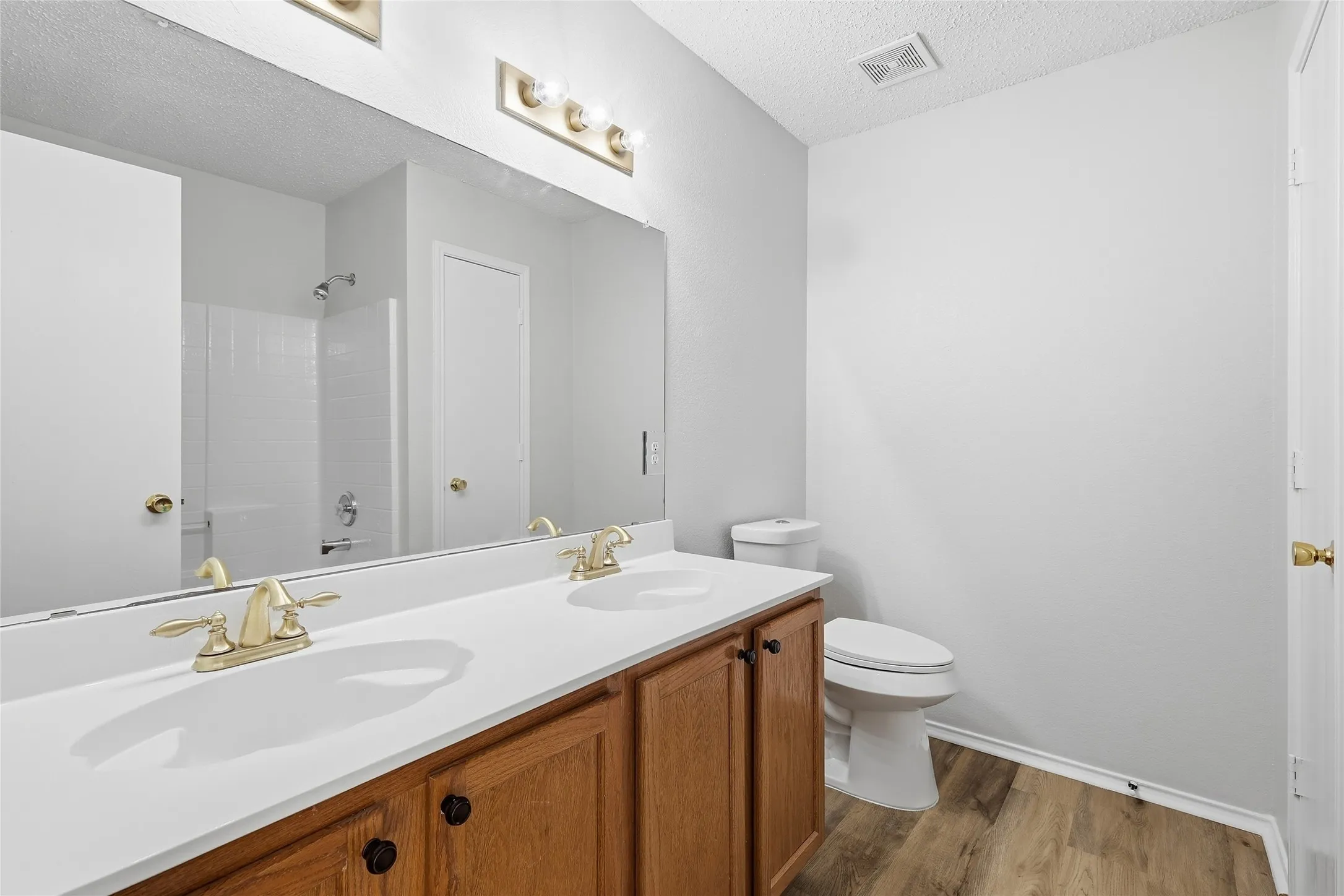 Dual vanity with brass-finish fixtures and wood-finish cabinetry, white countertops, full-width mirror, wood-look flooring, and a white toilet