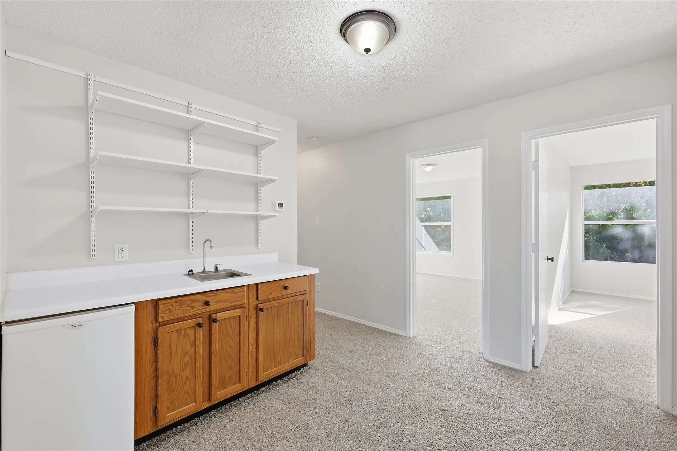 Built-in wooden cabinetry, white countertop, stainless steel sink with faucet, white wall-mounted shelving, and light gray carpeted flooring