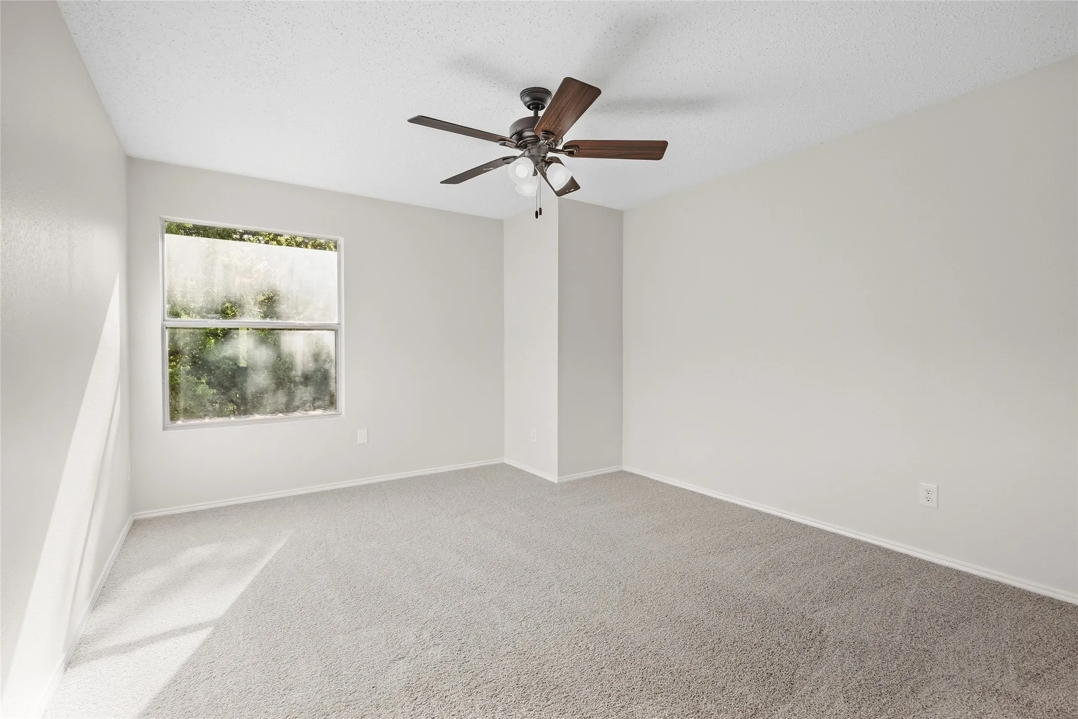 Spacious room featuring neutral carpeting, light-colored walls, a ceiling fan with integrated lighting, and natural light from a window