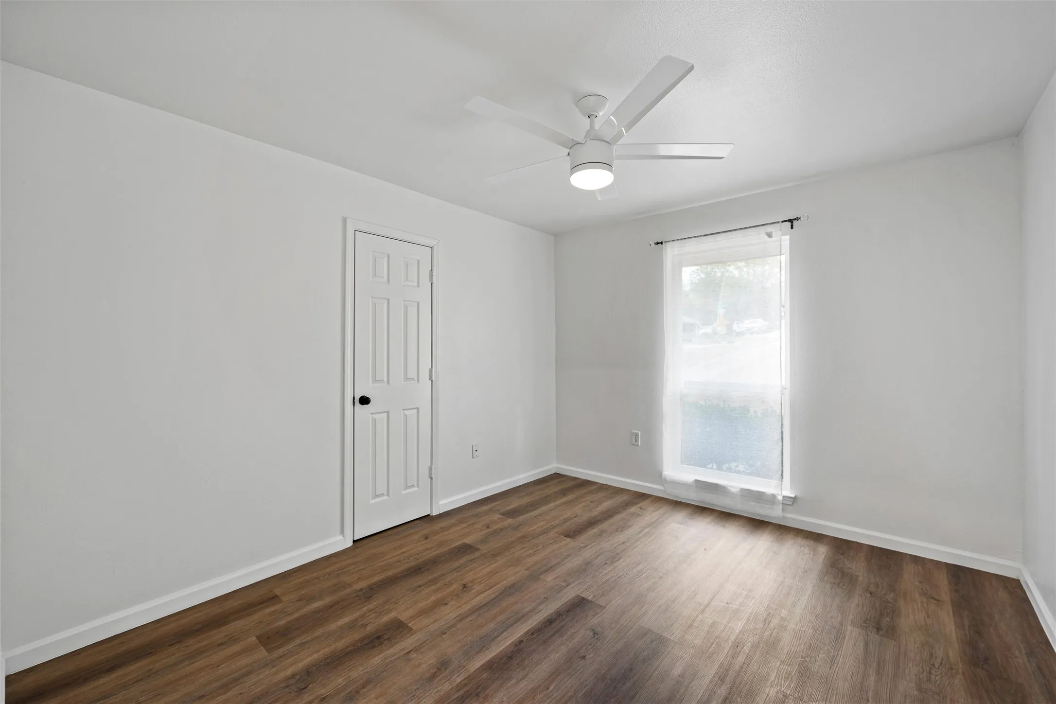 Wood-finish flooring, white paneled door, modern ceiling fan with integrated lighting, and a single window with curtain rod