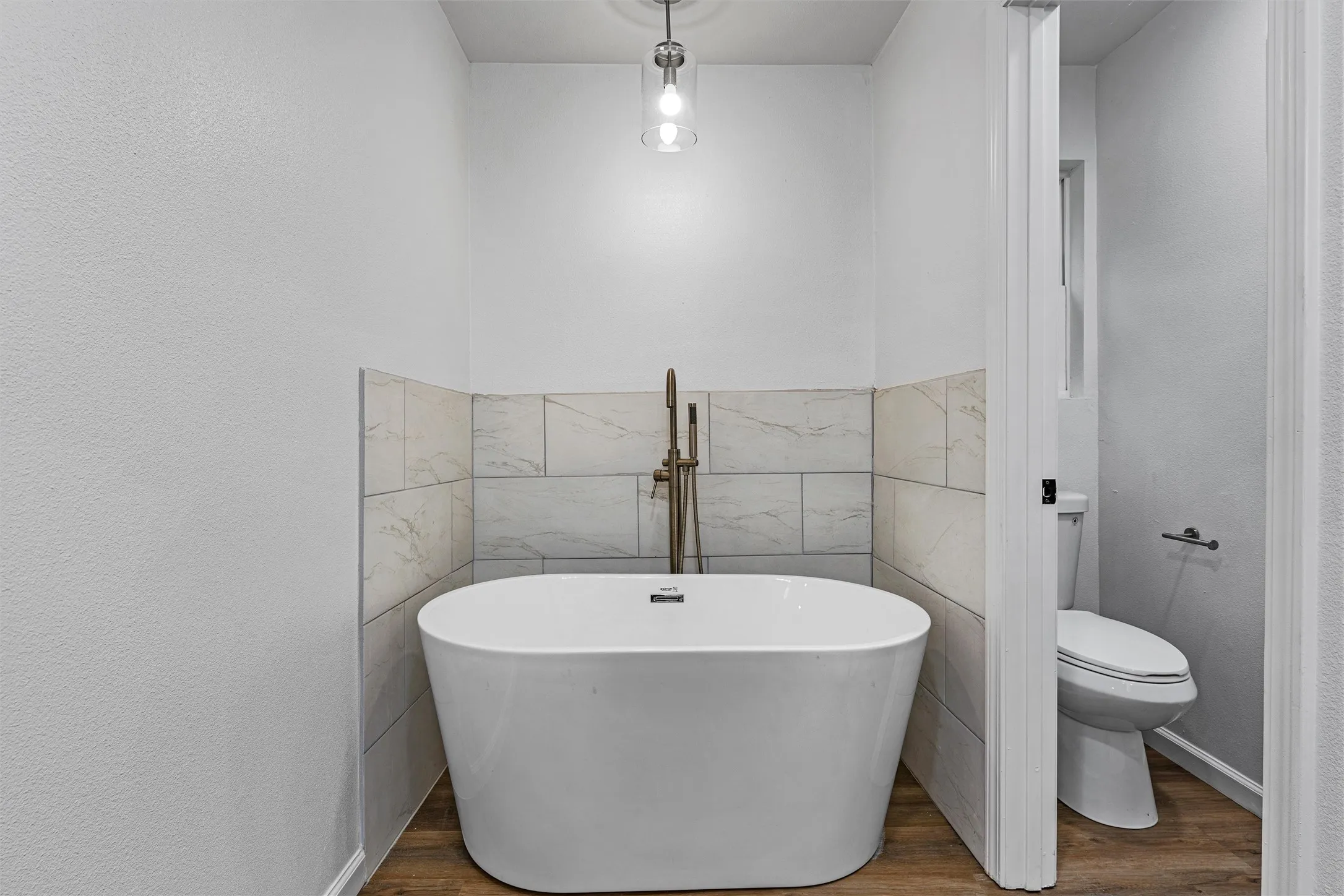Freestanding soaking tub with floor-mounted faucet, tile wainscoting, wood-finish flooring, and a contemporary pendant light fixture
