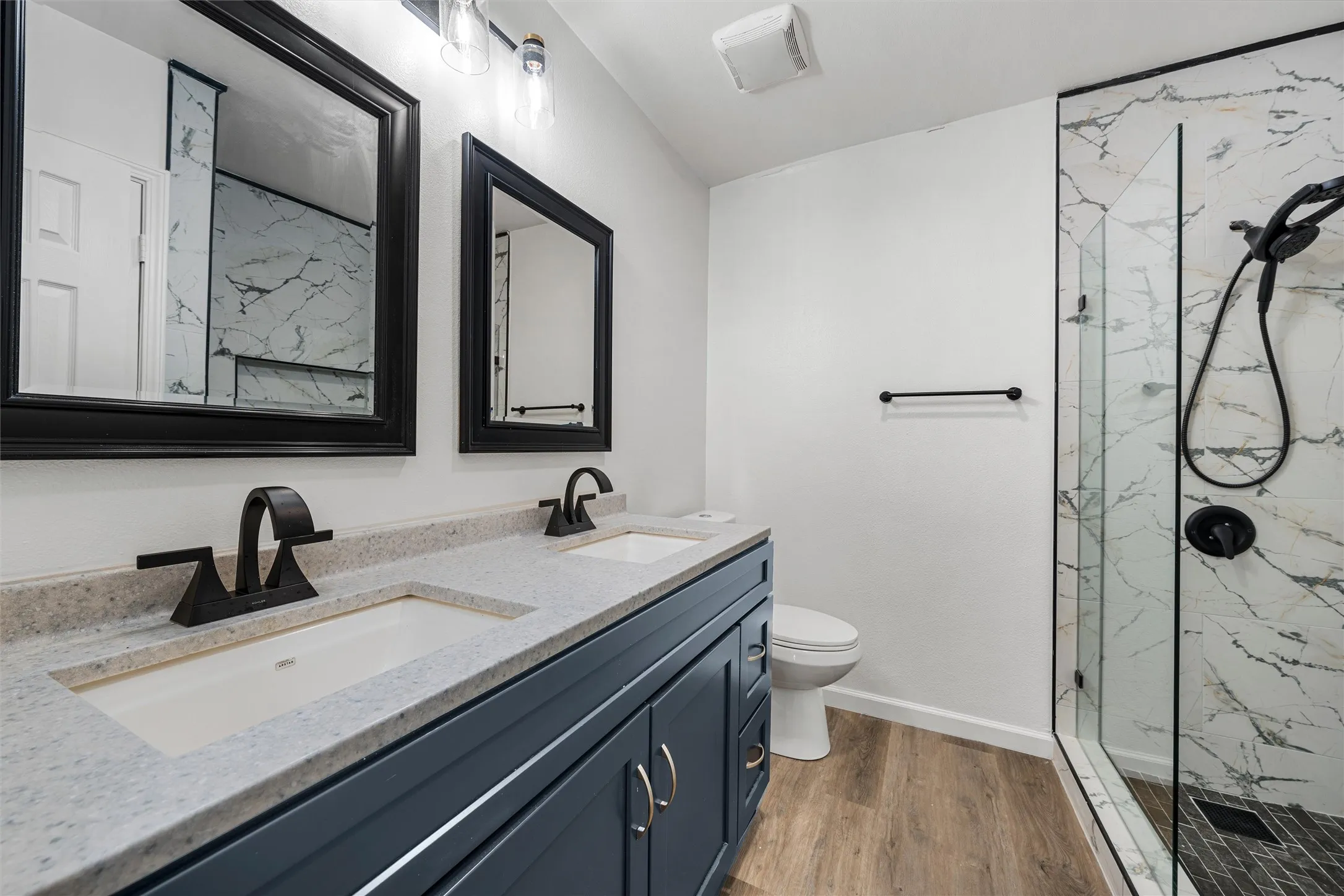 Dual vanity with integrated sinks and dark blue cabinetry, quartz-finish countertop, black-framed mirrors, and a glass-enclosed shower with a marble-patterned tile surround