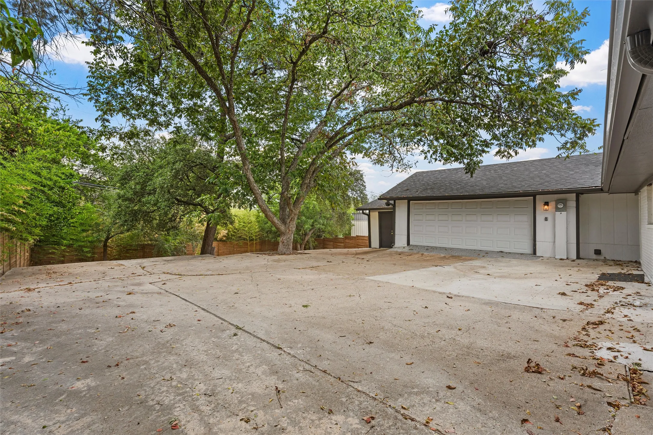 Expansive concrete driveway leading to a multi-car garage with a panel door, accompanied by mature trees and a privacy fence
