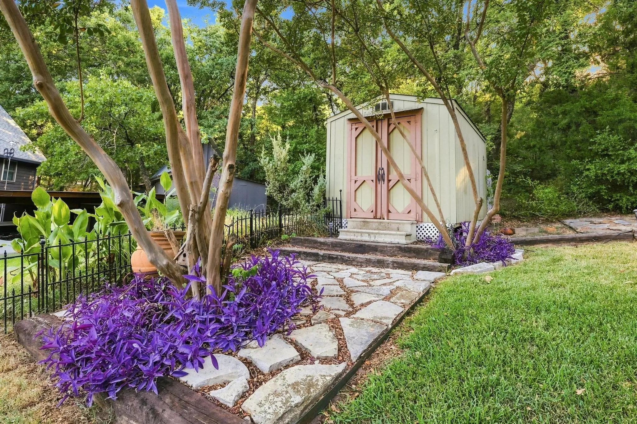 Flagstone pathway leading to a detached storage shed with double doors, surrounded by vibrant purple ground cover and mature trees