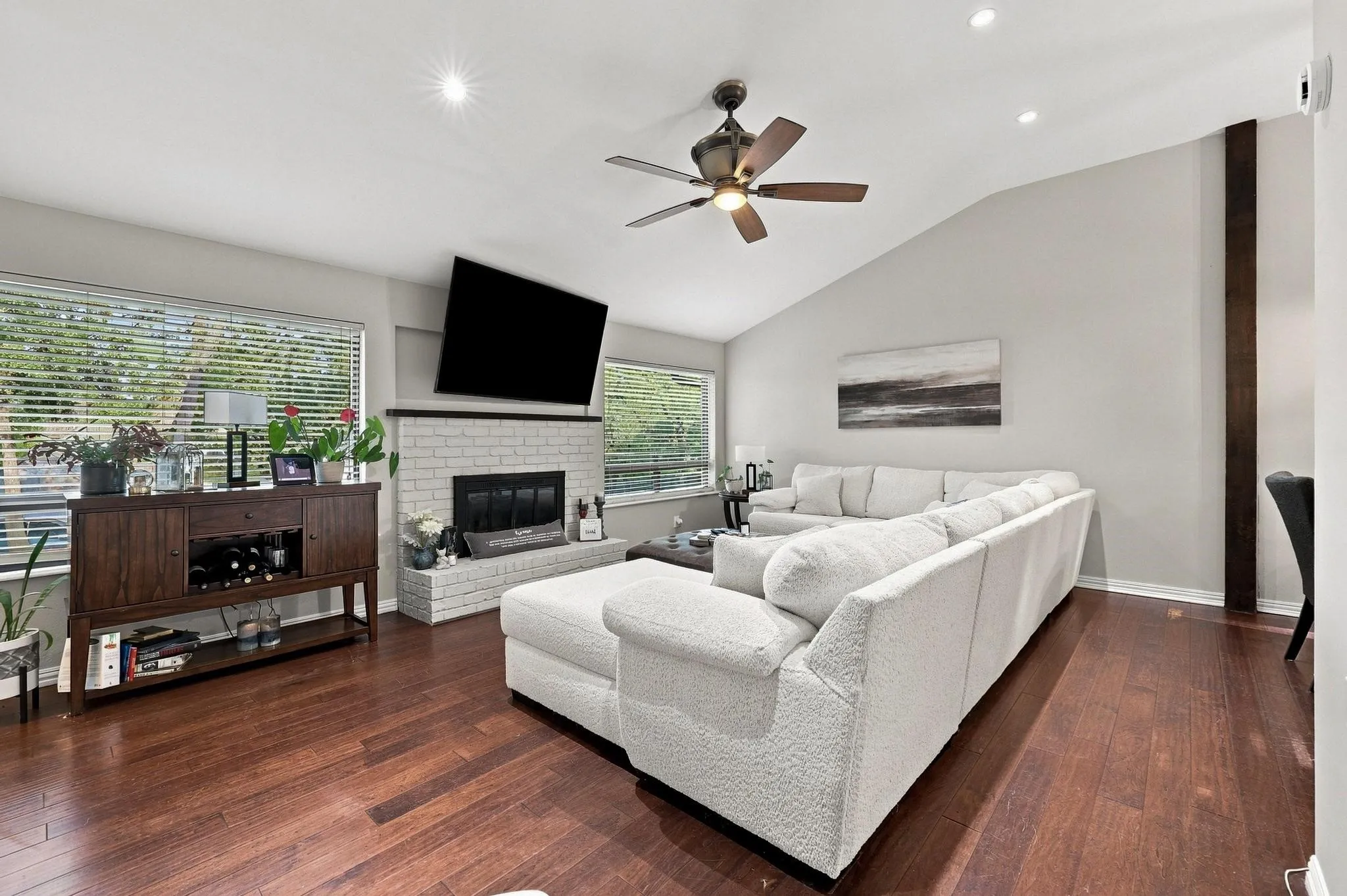 Vaulted ceiling with recessed lighting, wood-finish flooring, brick-surround fireplace, ceiling fan, and large windows with blinds