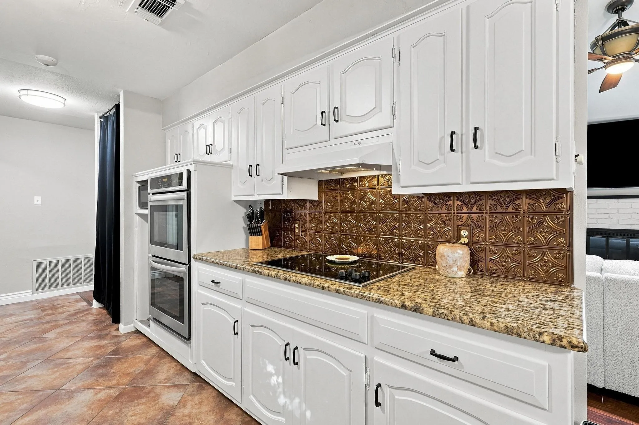 White cabinetry with black hardware, double wall ovens, built-in cooktop, patterned backsplash, and textured tile flooring