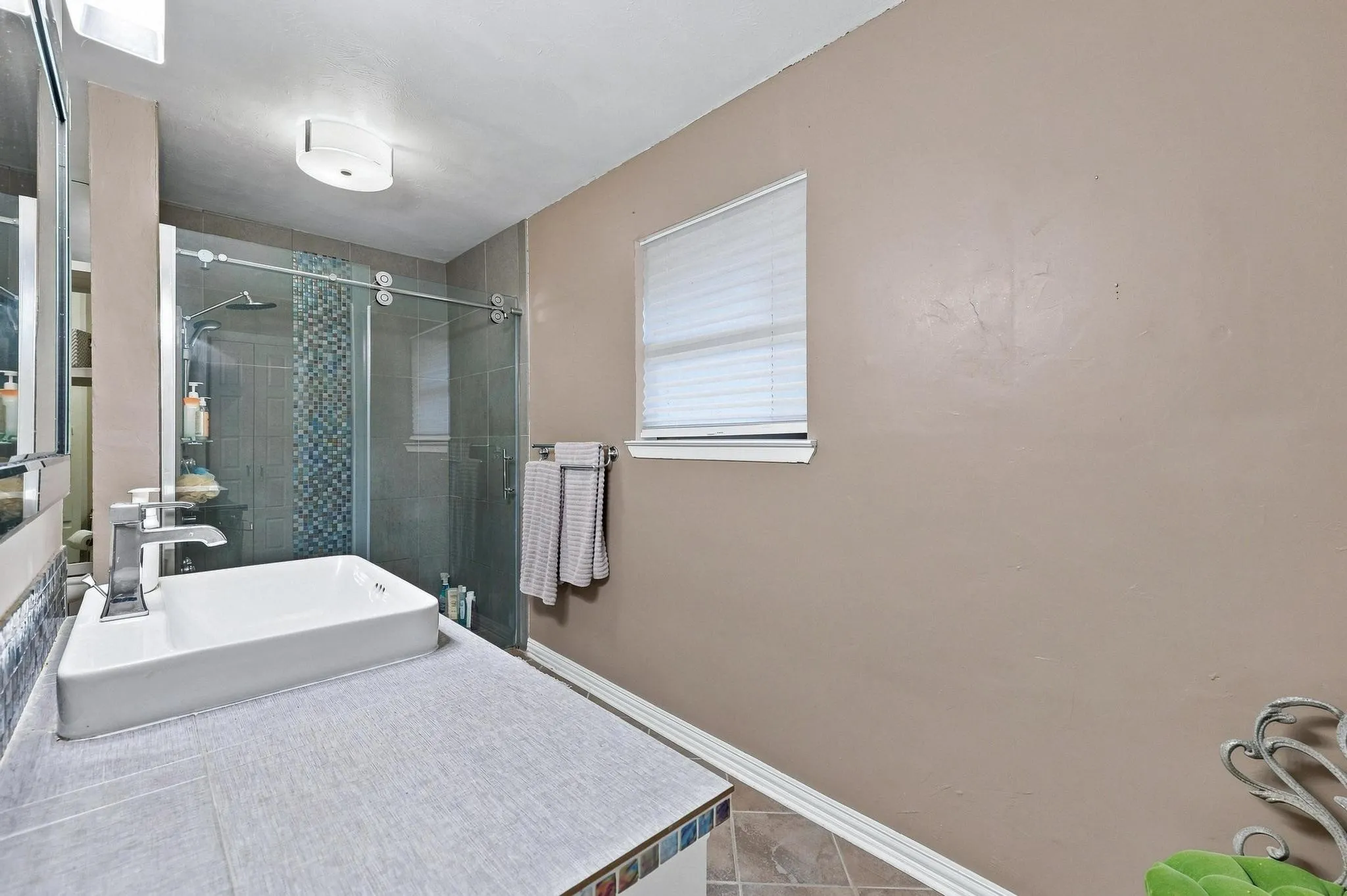 Rectangular vessel sink with a polished chrome faucet, tiled countertop, frameless glass shower enclosure, and mosaic tile shower accent