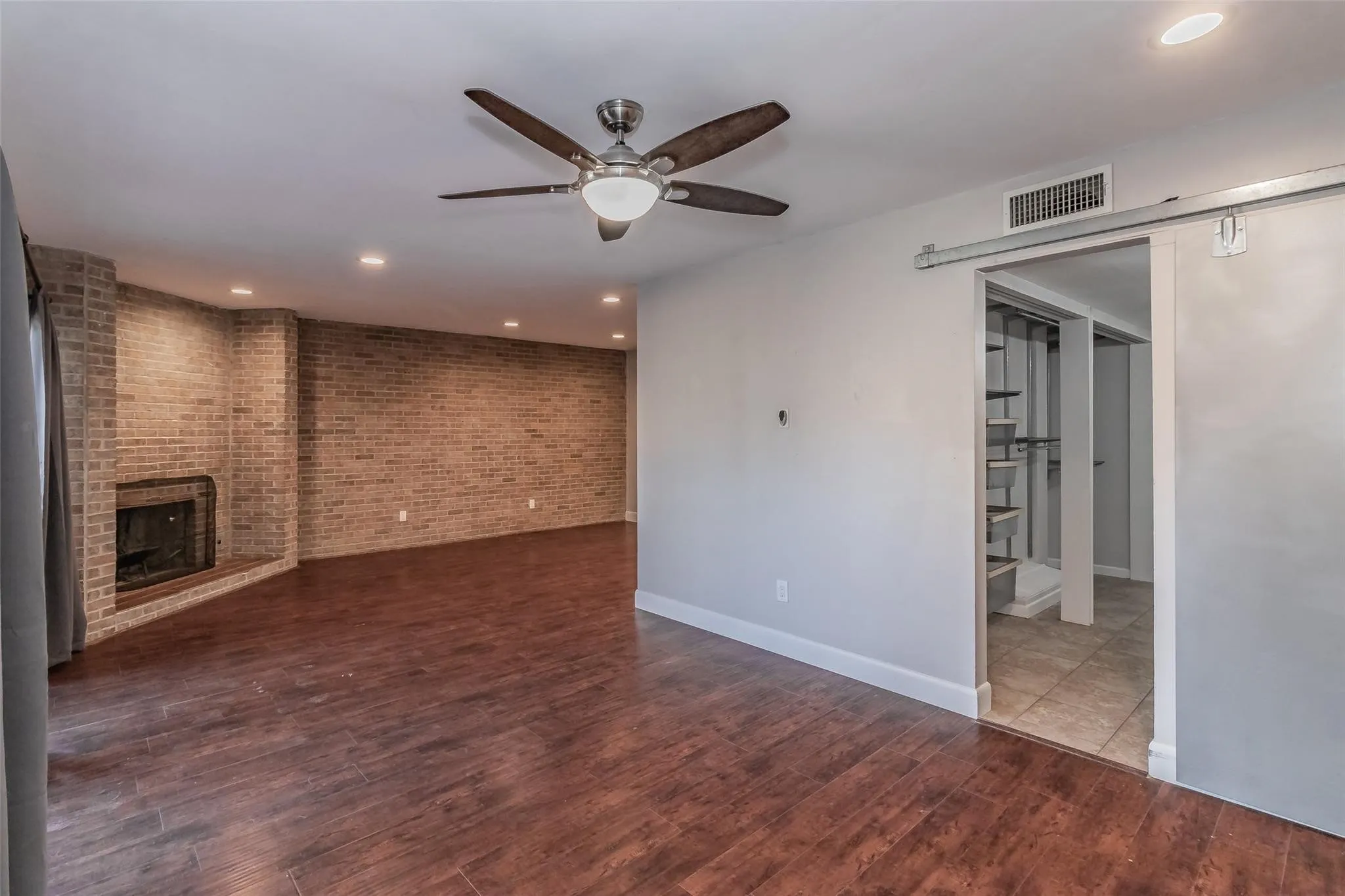 Wood-finish flooring, exposed brick wall with fireplace, recessed lighting, ceiling fan, and barn door entry to an additional space