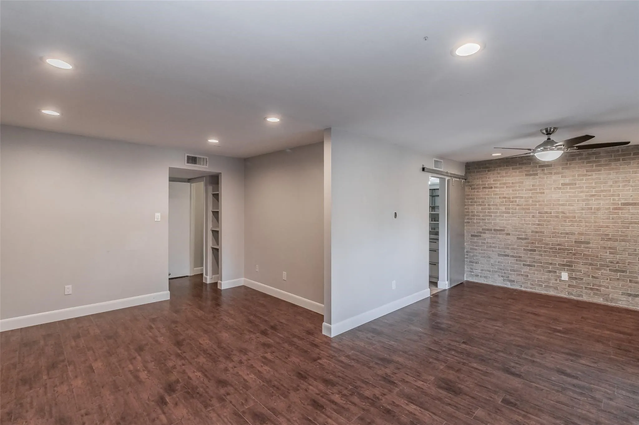 Wood-finish flooring throughout, recessed ceiling lighting, a ceiling fan with integrated lighting, a brick-finish accent wall, and a barn door leading to an interior space with built-in shelving