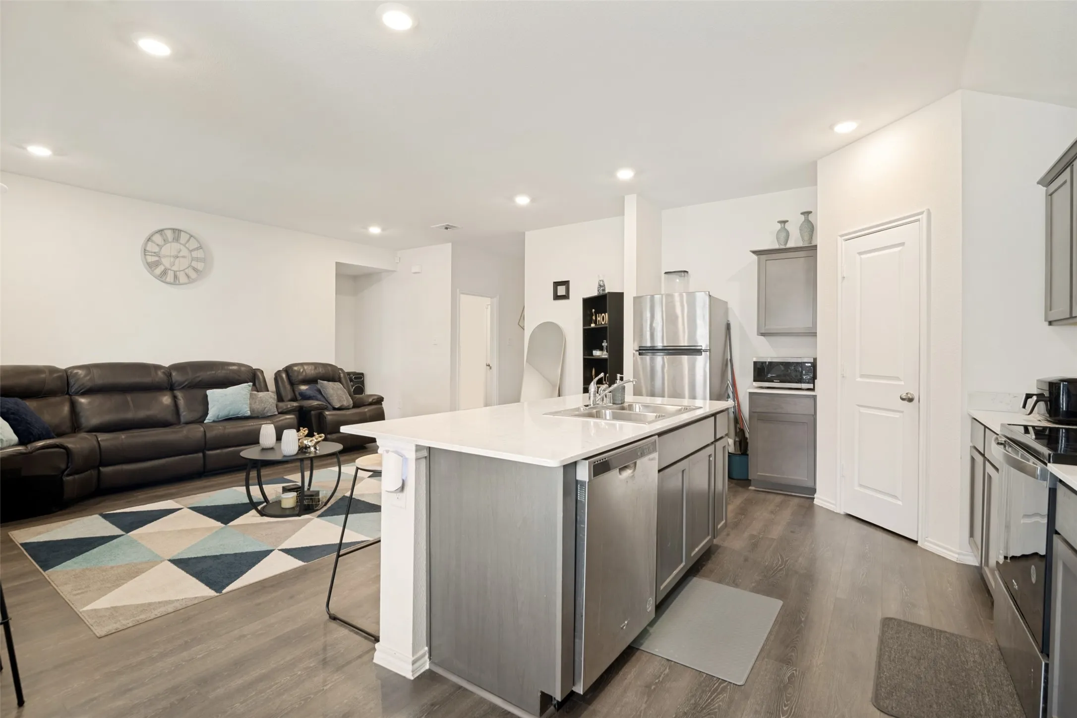 Open-concept living area featuring wood-finish flooring, recessed lighting, a kitchen island with an integrated sink, stainless steel appliances, and grey cabinetry