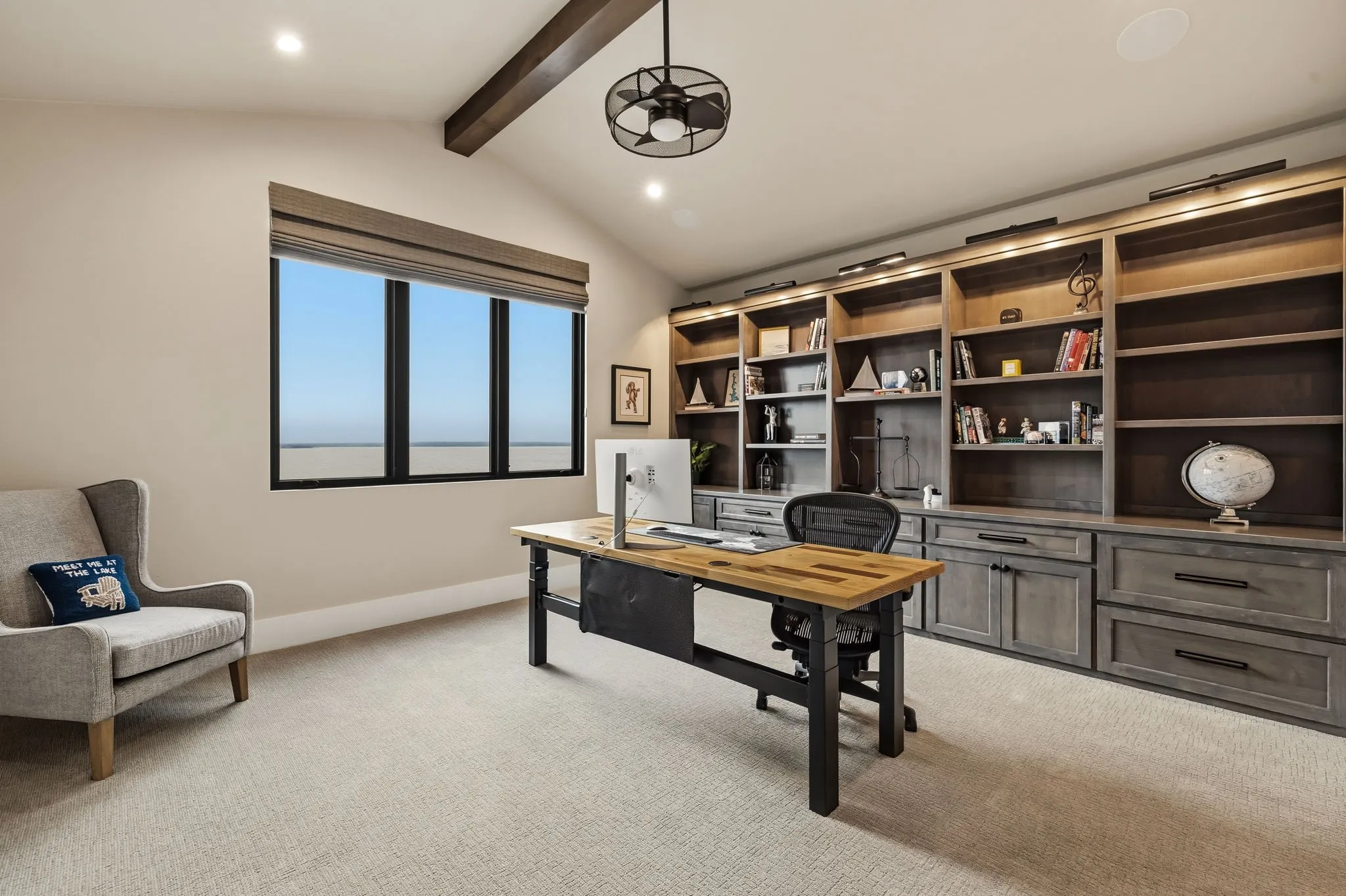 Vaulted ceiling with exposed wood beam, built-in shelving with integrated lighting, wall-mounted window treatment, and neutral-toned carpeting