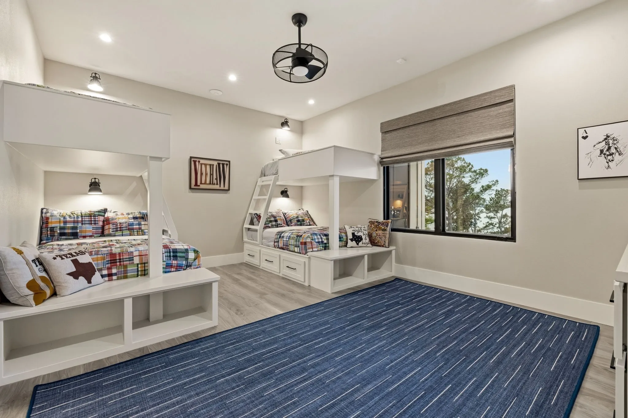 Built-in bunk beds with integrated storage, wood-finish flooring, a large window with a roller shade, and a ceiling fan