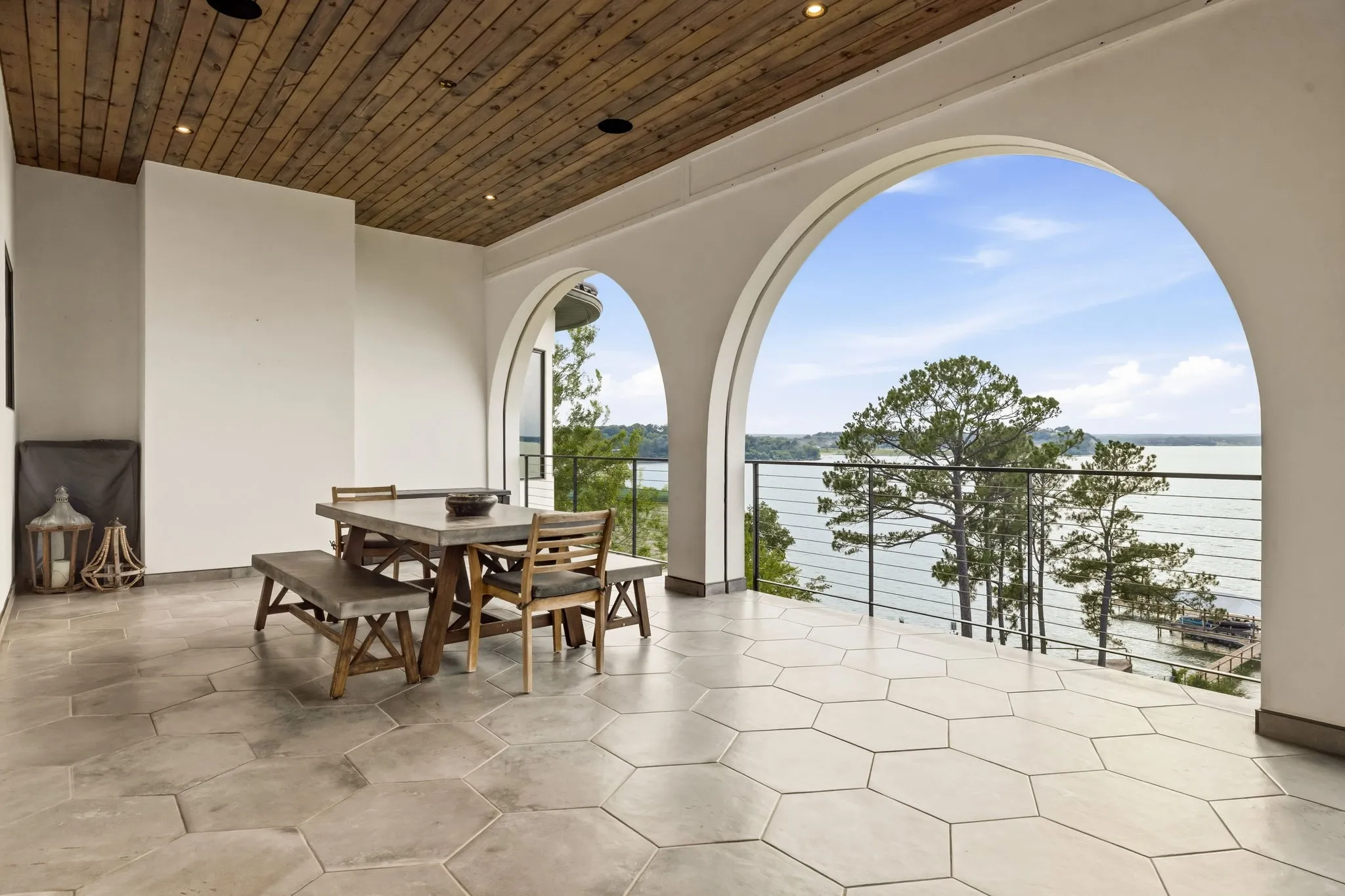 Covered patio with a wood-plank ceiling, hexagonal floor tiles, and archways framing a water backdrop with mature trees