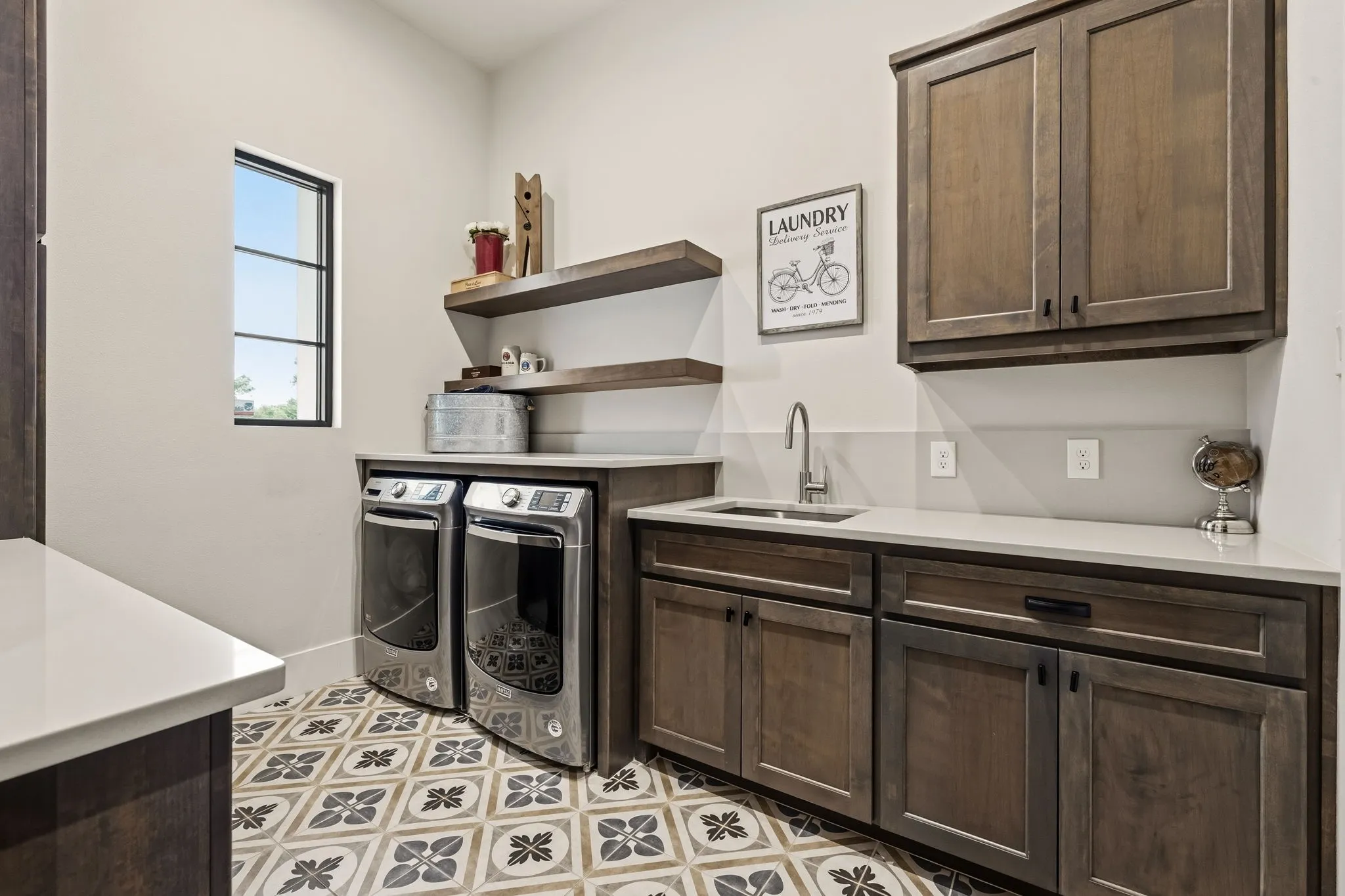 Dedicated laundry room featuring dual front-load washers and dryers, quartz countertop with undermount sink and chrome faucet, dark wood cabinetry with ample storage, floating wood shelves, and decorative tile flooring in a geometric pattern