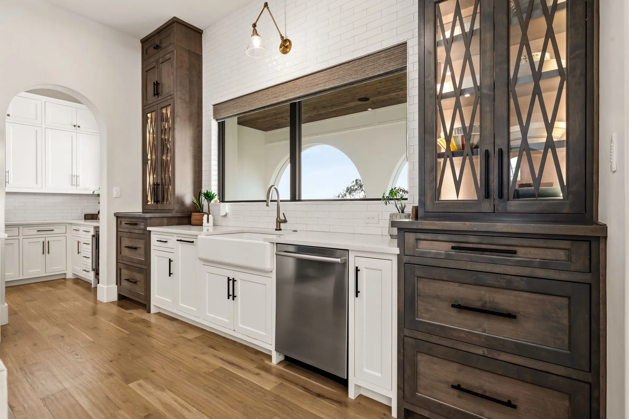 Kitchen with wood-finish flooring, white cabinetry, a farmhouse sink, stainless steel dishwasher, and dark wood-finish built-in storage with glass-front display cabinets