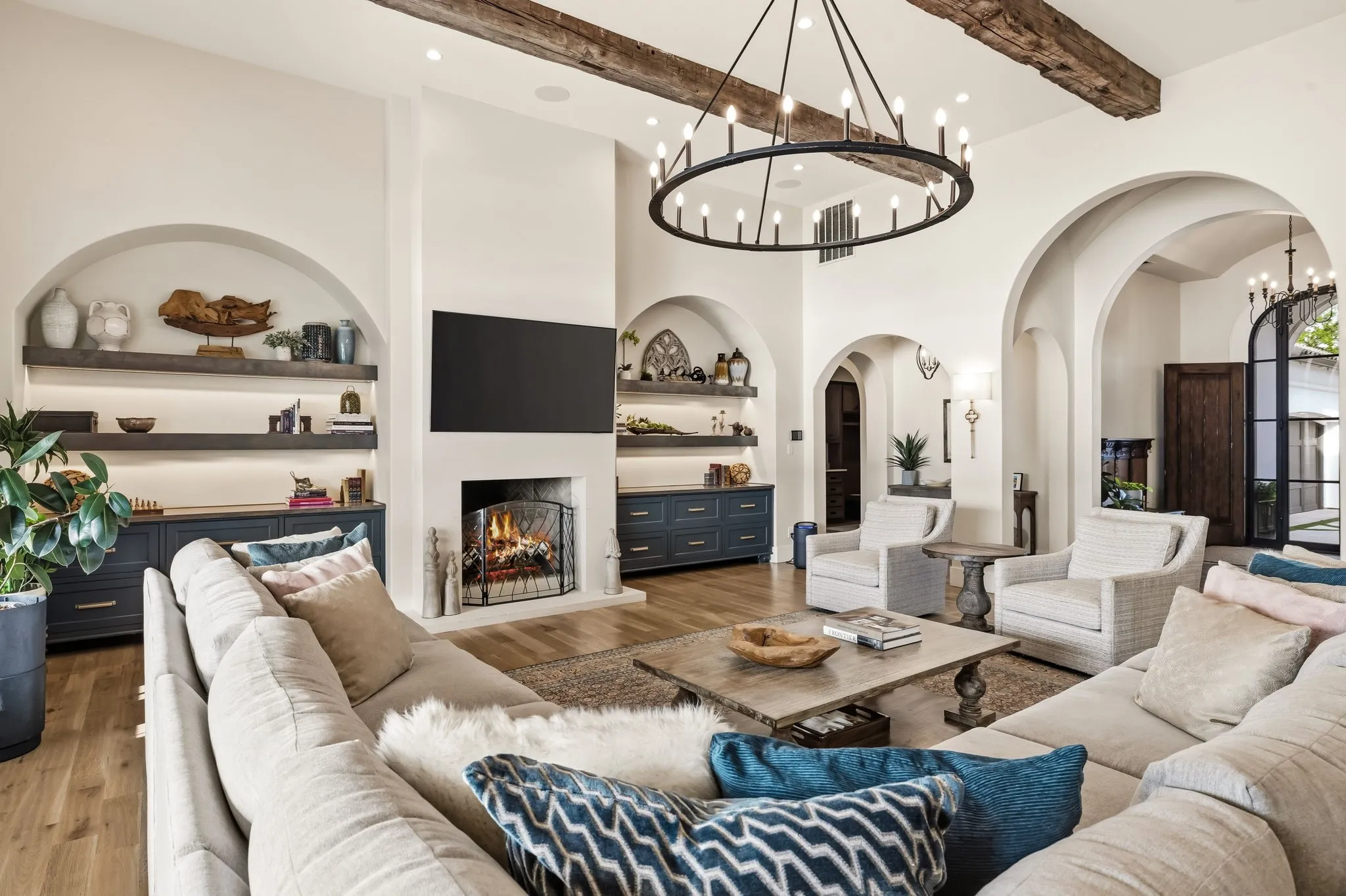 Vaulted ceiling with exposed wood beams, multiple arched doorways, wood-finish flooring, built-in fireplace, and custom cabinetry with floating shelving