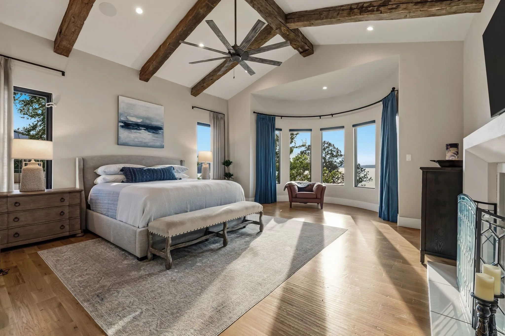 Vaulted ceiling with exposed wood beams, wood-finish flooring, a curved wall of windows, and an integrated ceiling fan
