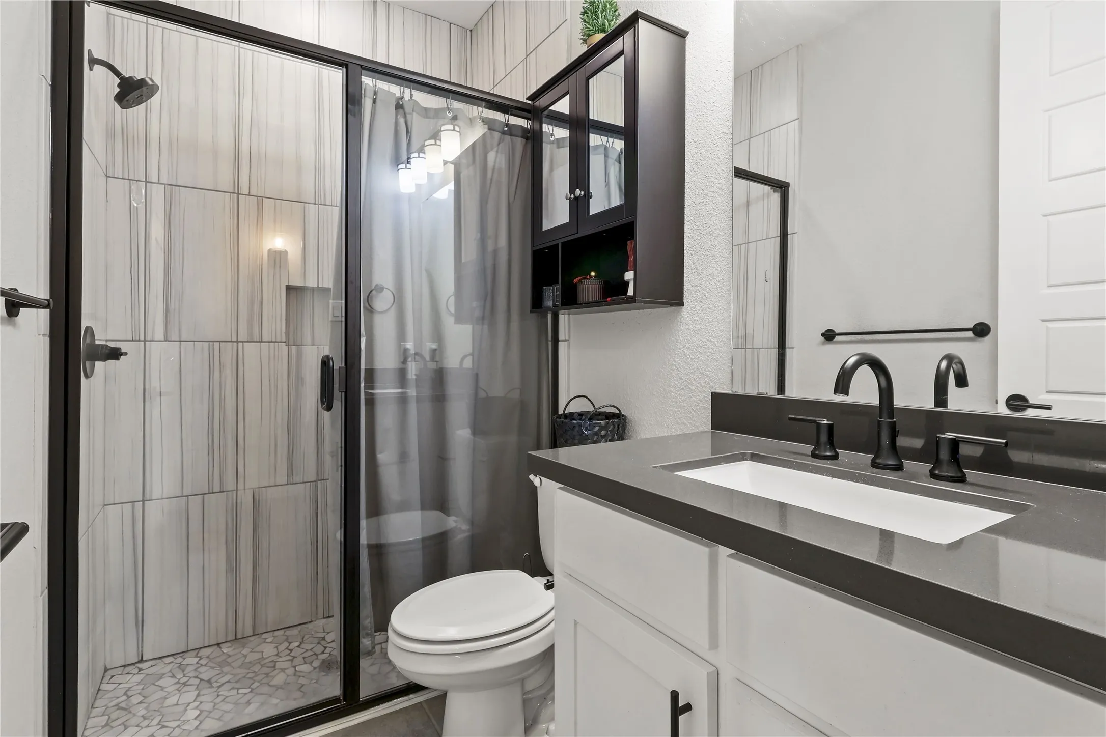 Shower with floor-to-ceiling vertically-stacked tile, glass enclosure with matte black framing, and pebble-style flooring
