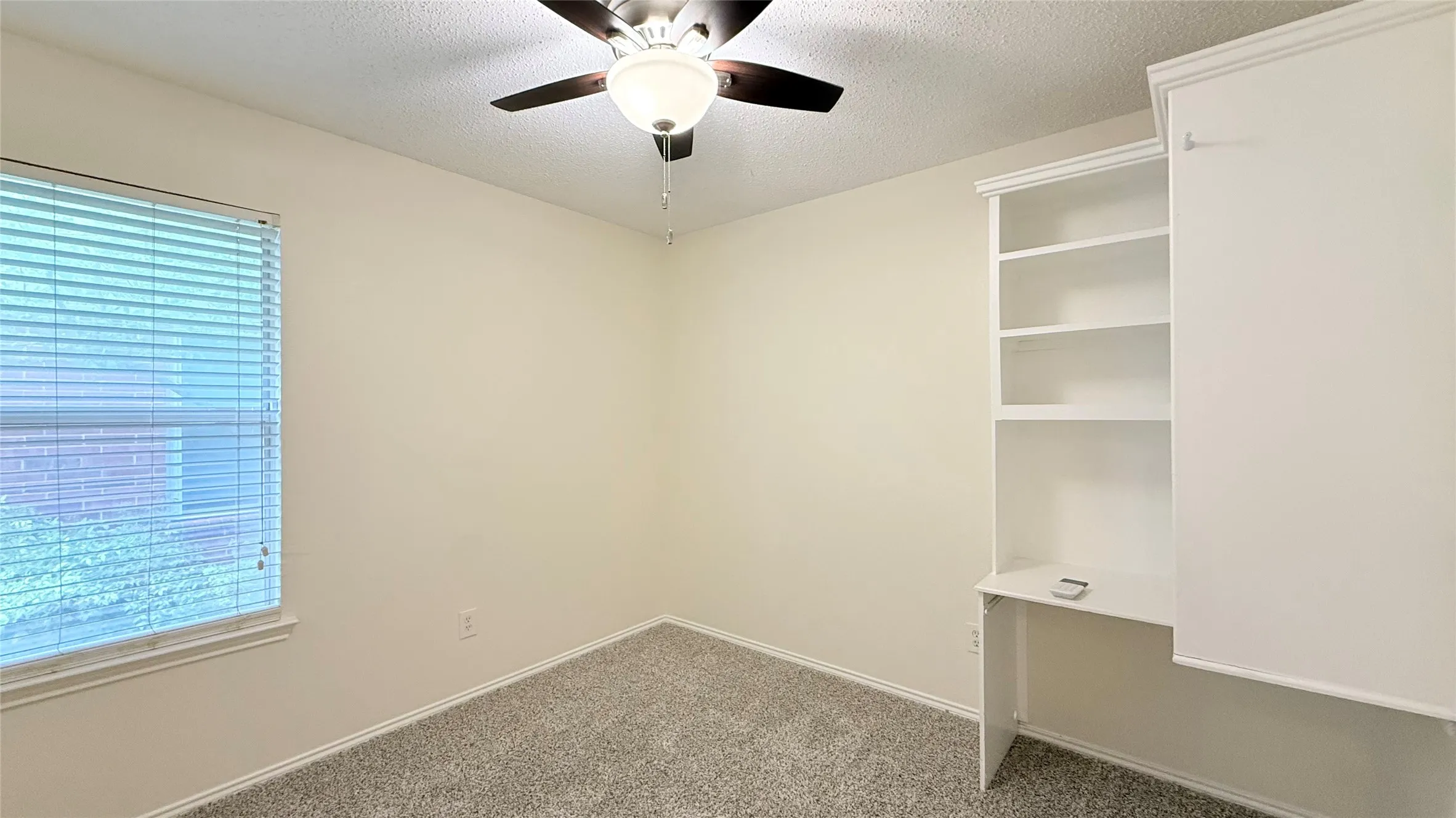 Neutral wall paint, textured ceiling, ceiling fan with integrated lighting, carpet flooring, and built-in shelving with desk space