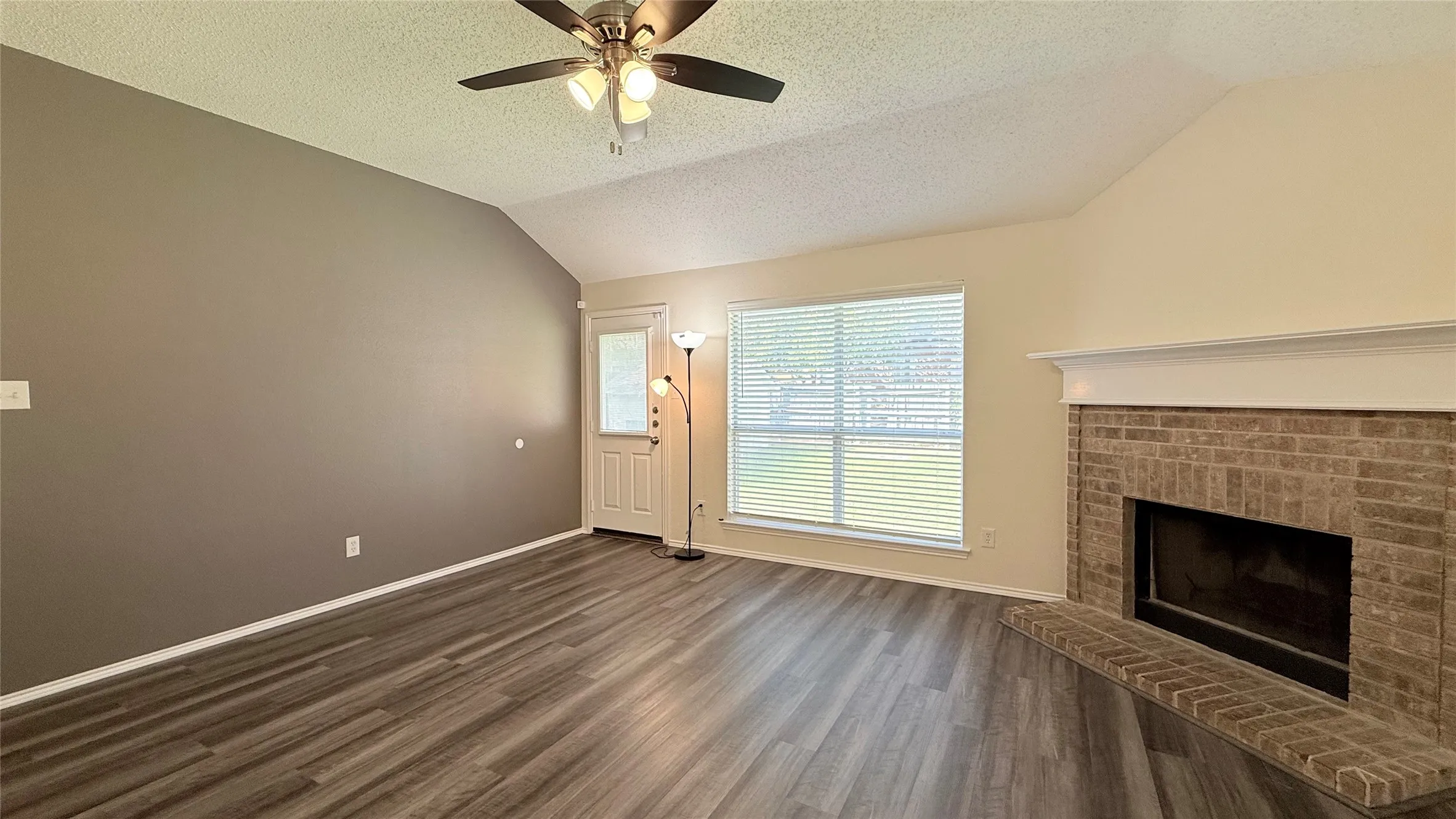 Vaulted ceilings, wood-finish flooring, brick fireplace with mantel, ceiling fan, and large window