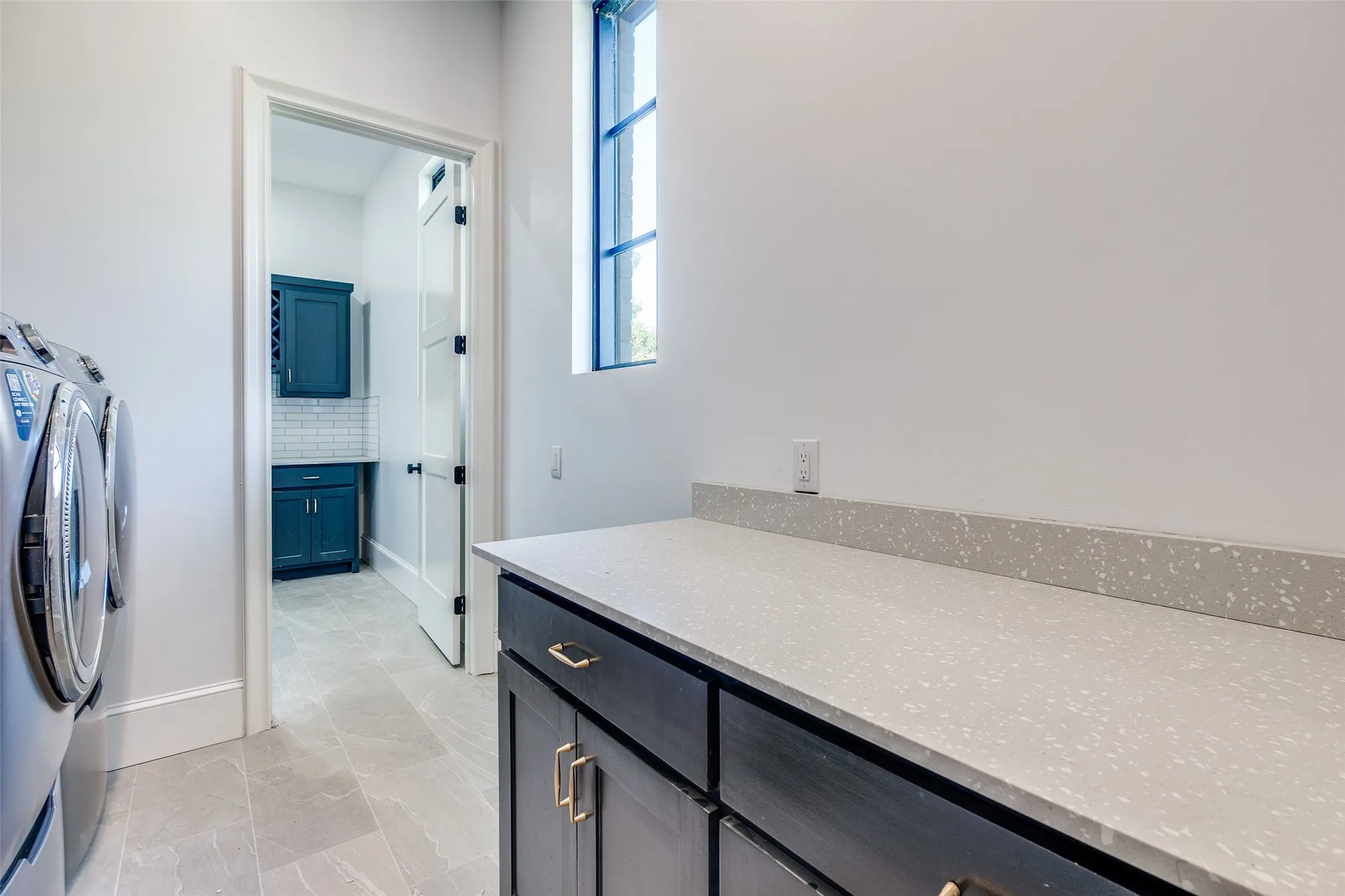 Light-toned tile flooring, built-in cabinetry with gold-toned hardware, solid surface countertop, and a tall window