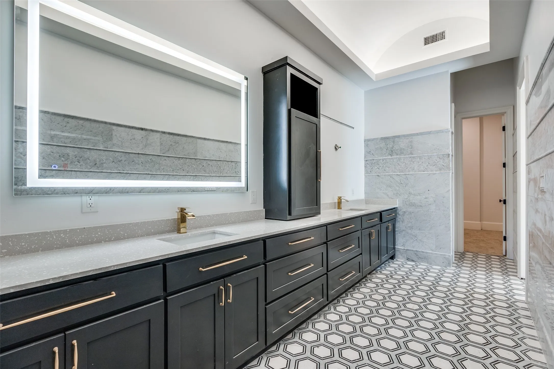 Dual vanity with integrated sinks, illuminated mirror, dark cabinetry with brass hardware, and geometric pattern tile flooring