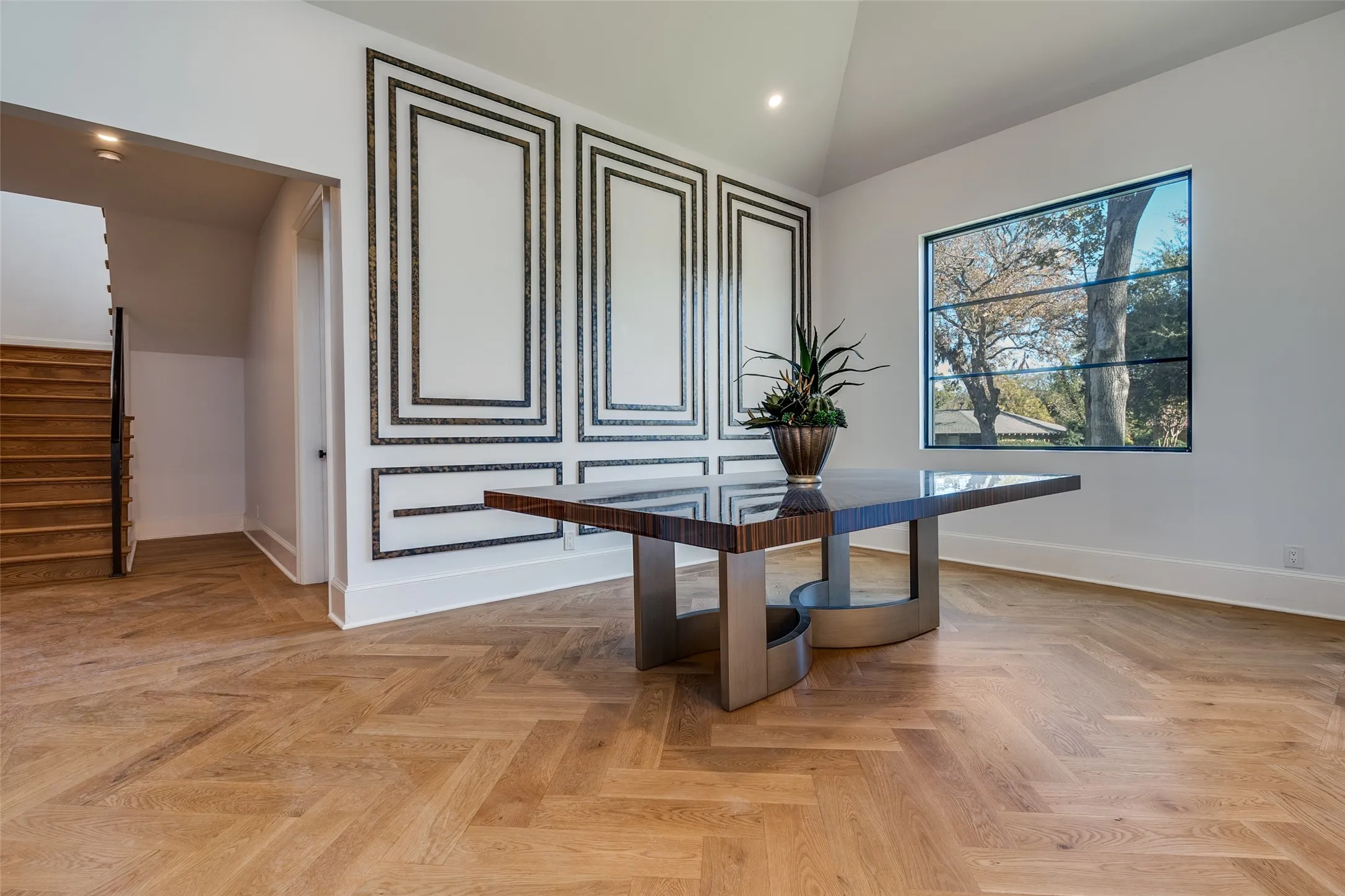 Herringbone wood-finish flooring, vaulted ceiling, built-in wall paneling with decorative trim, and a prominent window