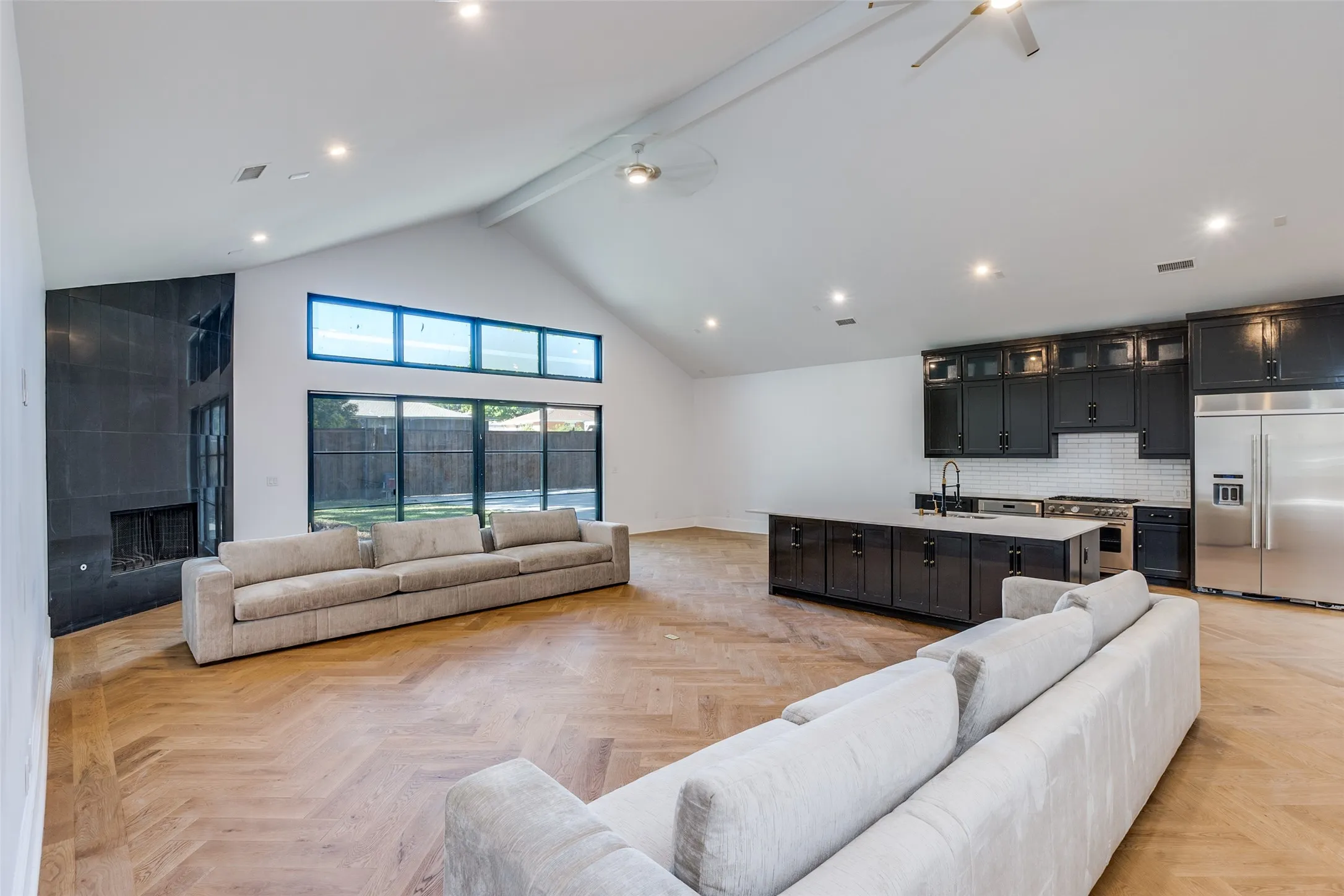 Vaulted ceiling with recessed lighting, wood-finish flooring, full height tiled fireplace, black framed windows, and a kitchen island with dark cabinetry
