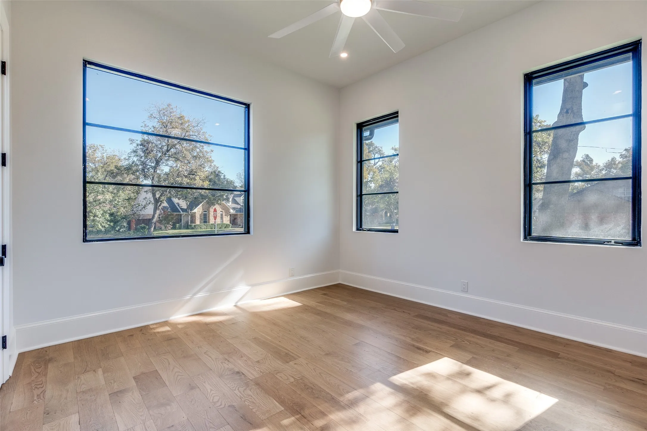 Wood-finish flooring, multiple black-framed windows, white walls, recessed lighting, and a ceiling fan