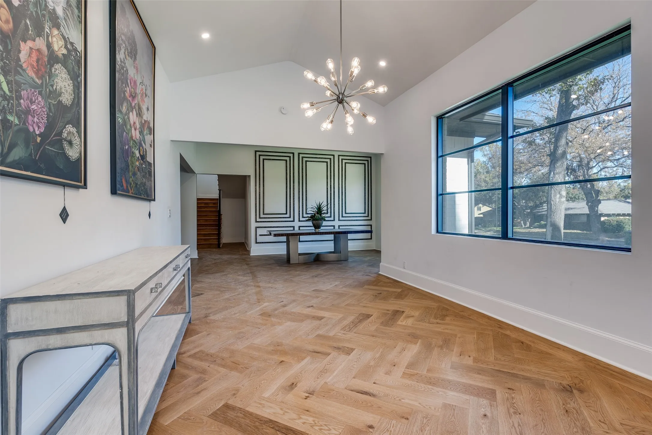Vaulted ceiling with a contemporary chandelier, hardwood flooring in a herringbone pattern, large gridded window, and decorative wall paneling