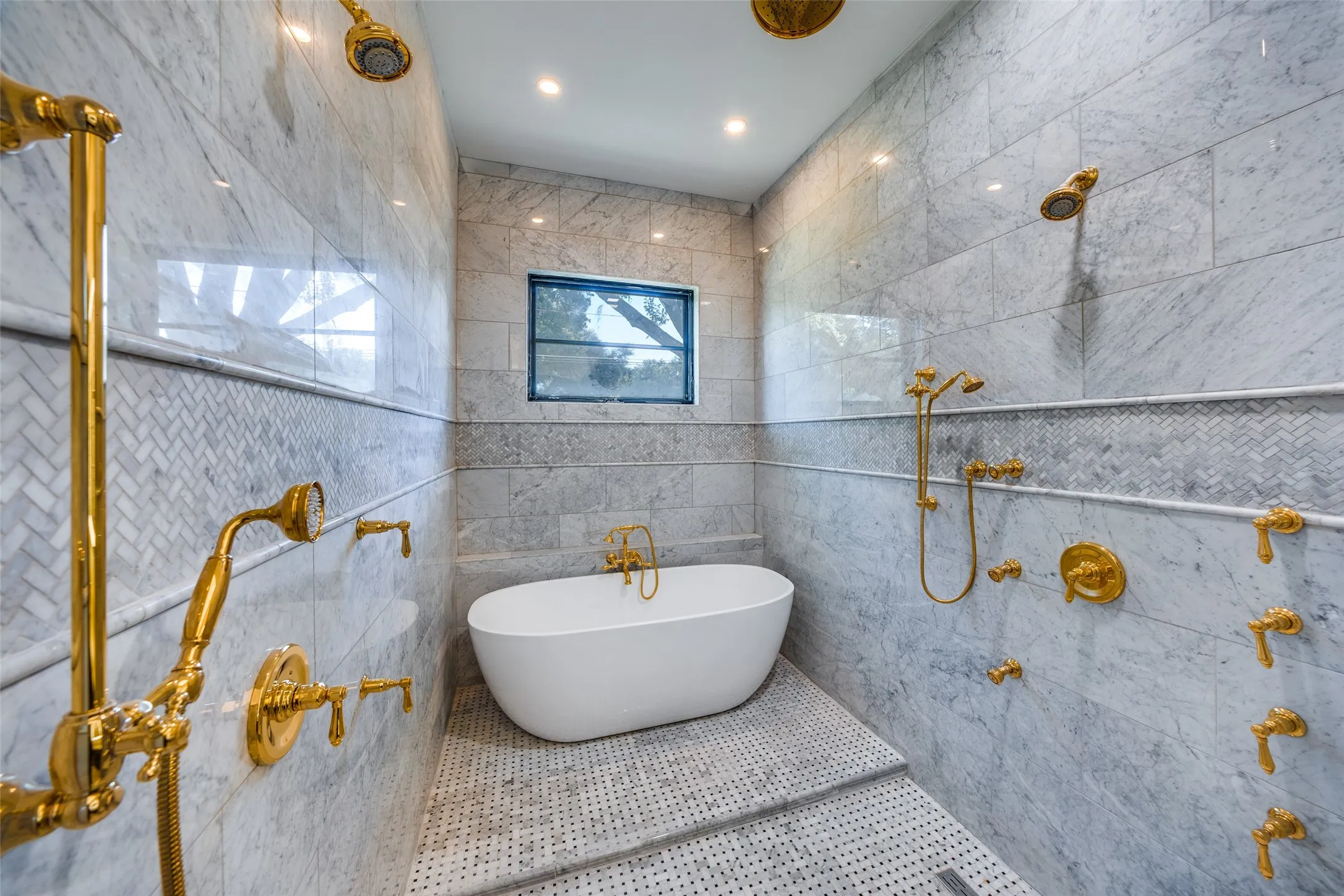 Freestanding bathtub within a large shower area, extensive wall tiling, herringbone tile accent, multiple shower heads, and gold-finish fixtures
