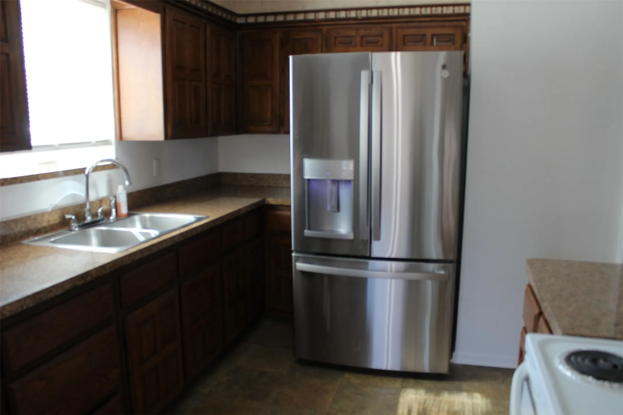 Dark wood-finish cabinetry, stainless steel French door refrigerator with ice/water dispenser, dual basin stainless steel sink with gooseneck faucet, light brown speckled countertops, and tile flooring