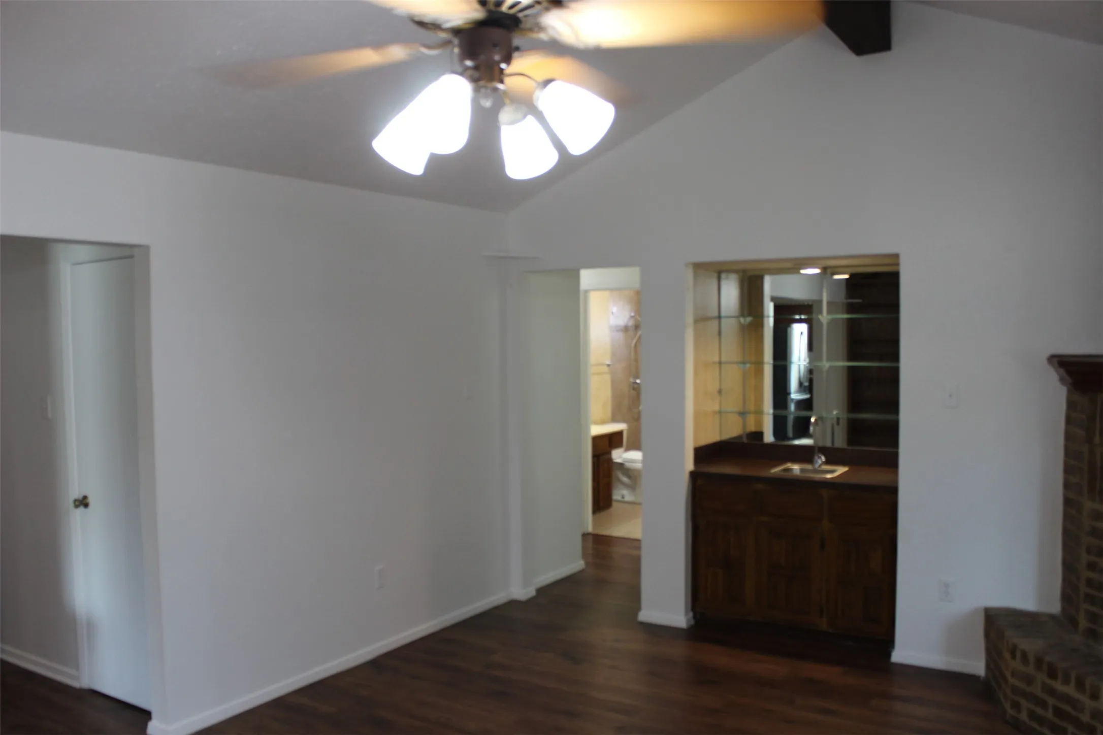 Vaulted ceiling, wood-finish flooring, built-in wet bar with mirrored backsplash and glass shelving, ceiling fan with integrated lighting, and a brick-faced fireplace surround