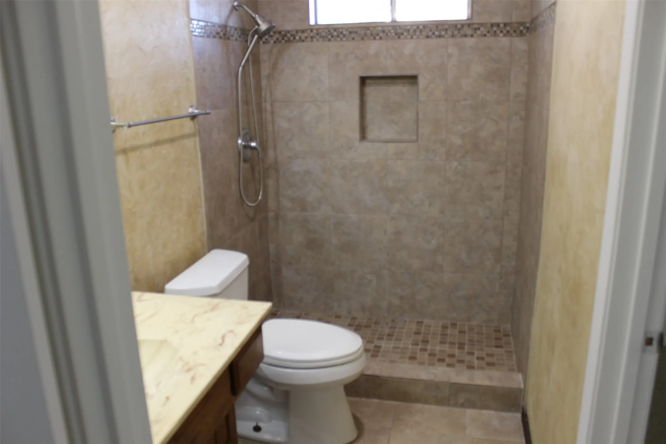 Tiled shower with an integrated niche and mosaic floor, alongside a vanity featuring a light-toned countertop and wood-finish cabinetry
