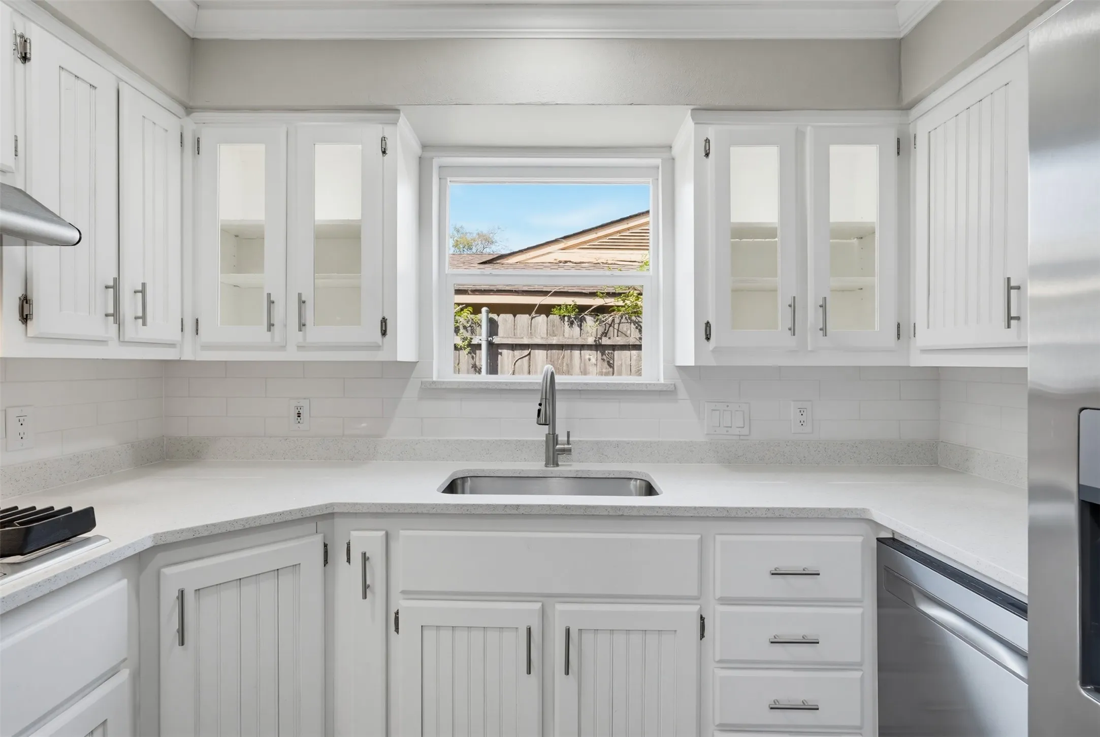 White cabinetry with glass-front uppers, stainless steel sink with a gooseneck faucet, light-toned countertops, and subway tile backsplash