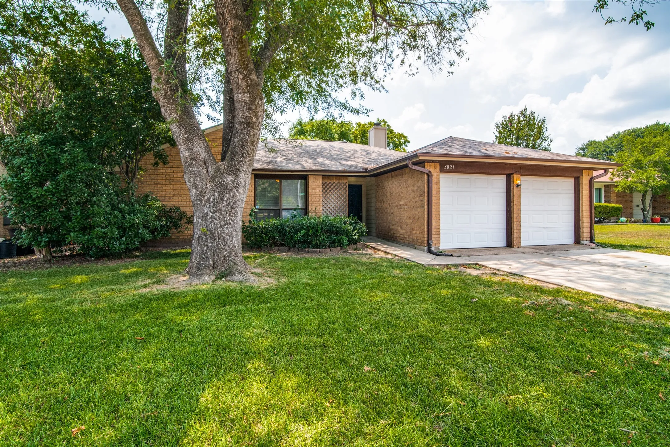 Brick exterior with a brown roof, featuring a two-car garage with white doors, a concrete driveway, and a mature tree in the front yard