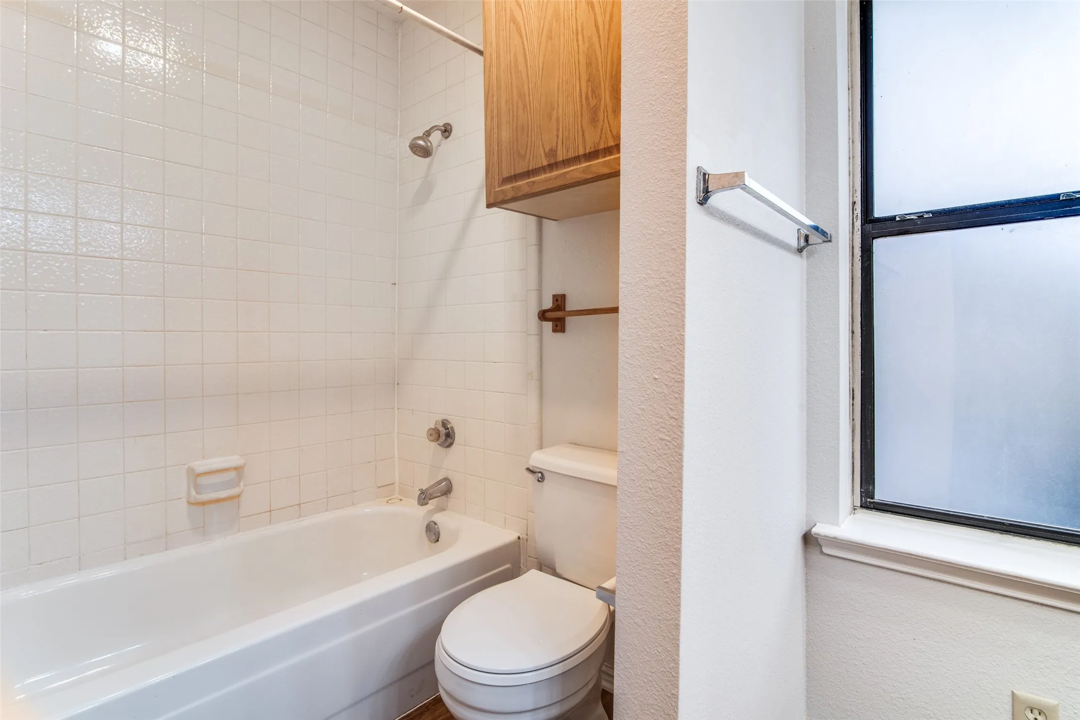 Bathtub with white tile surround, a shower fixture, and an overhead wooden cabinet