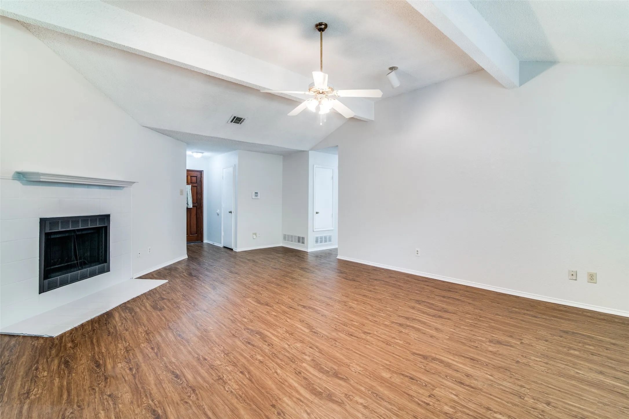 Vaulted ceiling with exposed beams, wood-finish flooring, white brick fireplace with dark tile surround, and ceiling fan with integrated lighting
