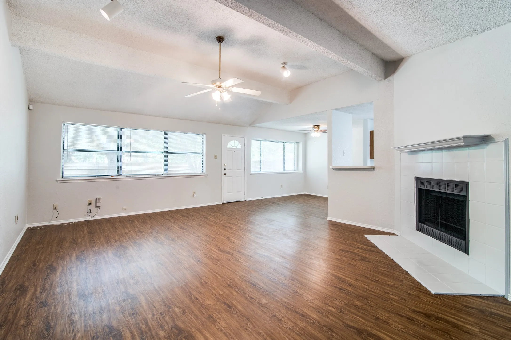 Wood-finish flooring throughout, vaulted ceiling with exposed beams, white tiled fireplace, multiple windows with blinds, and white entry door