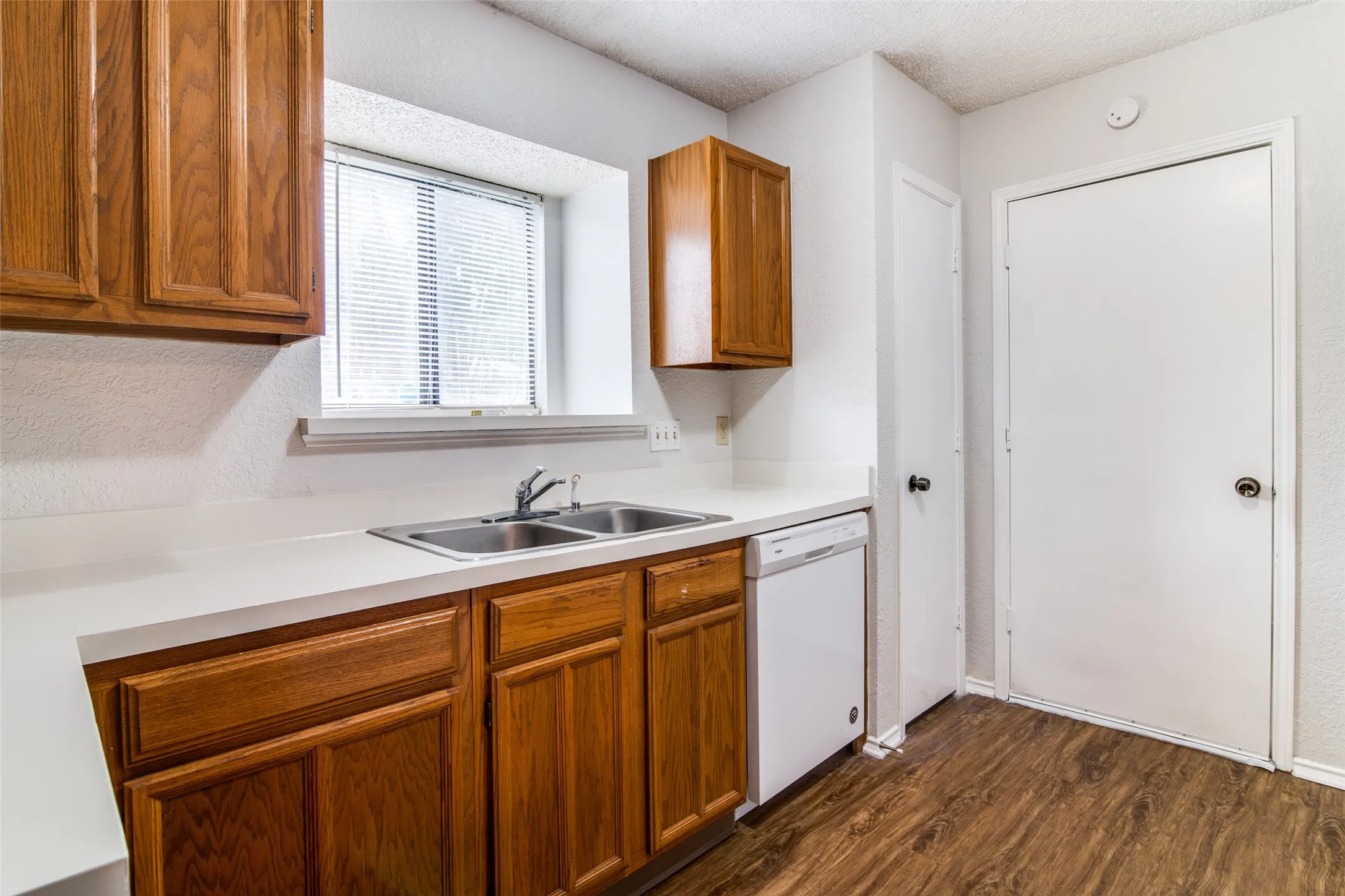 Kitchen featuring wood cabinetry, white countertops, a stainless steel double sink, white dishwasher, and wood-finish flooring
