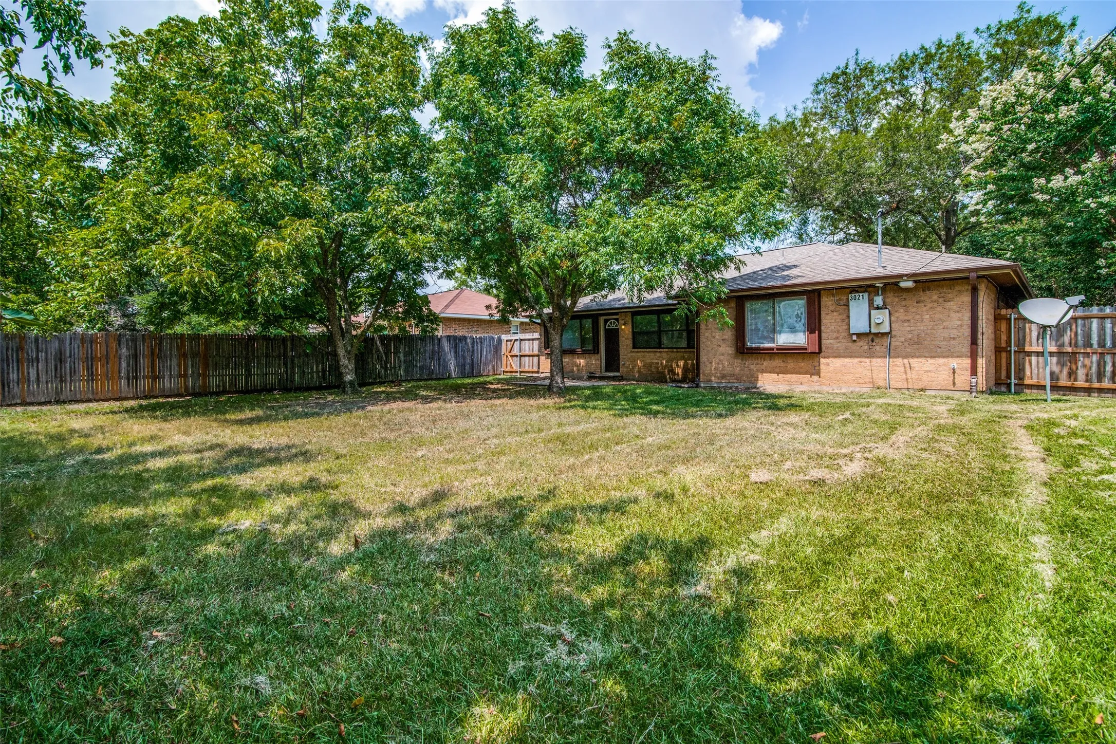 Expansive backyard featuring a privacy fence, mature trees, and a brick-clad exterior