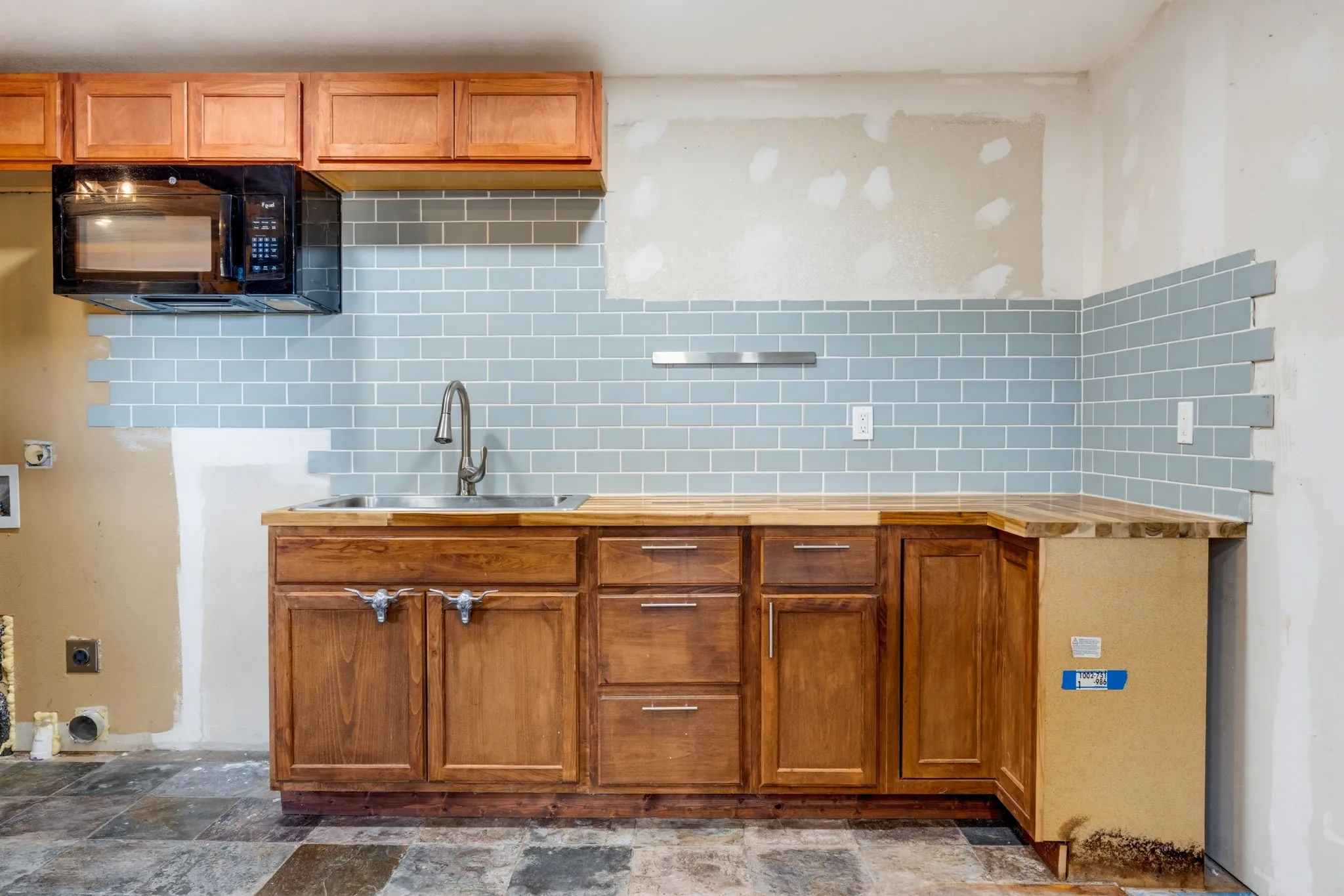 Wood-finish cabinetry with a built-in microwave, stainless steel sink, and light blue subway tile backsplash