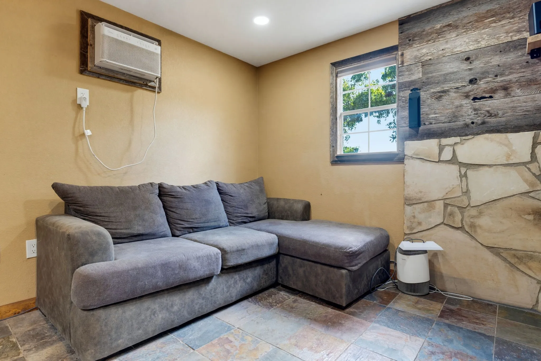 Recessed ceiling lighting, window-mounted air conditioning unit, rustic wood accent wall, stone-clad fireplace surround, and multi-toned slate-style flooring