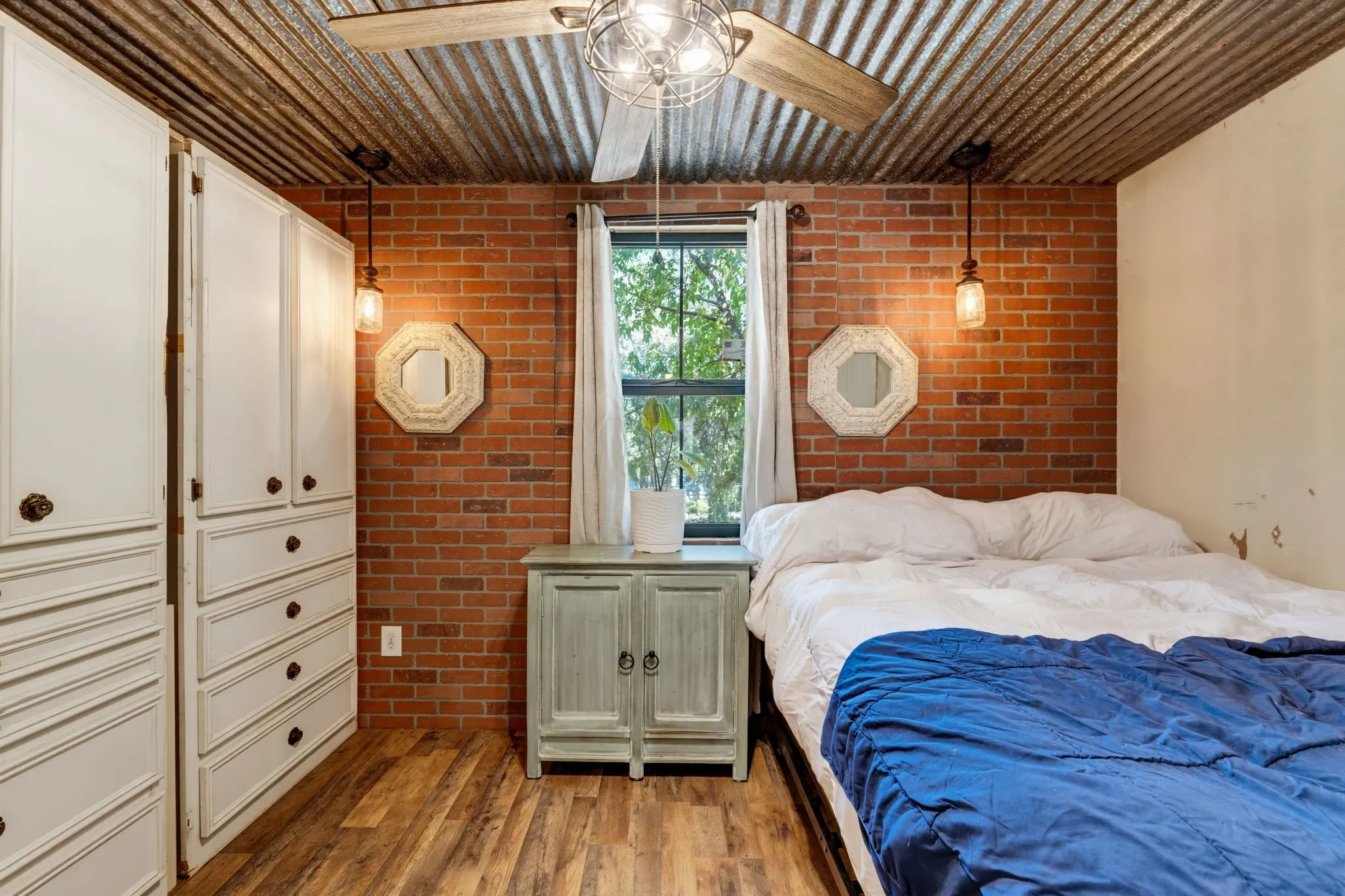 Corrugated metal ceiling, exposed brick accent wall, wood-finish flooring, built-in white cabinetry, and a ceiling fan with integrated lighting