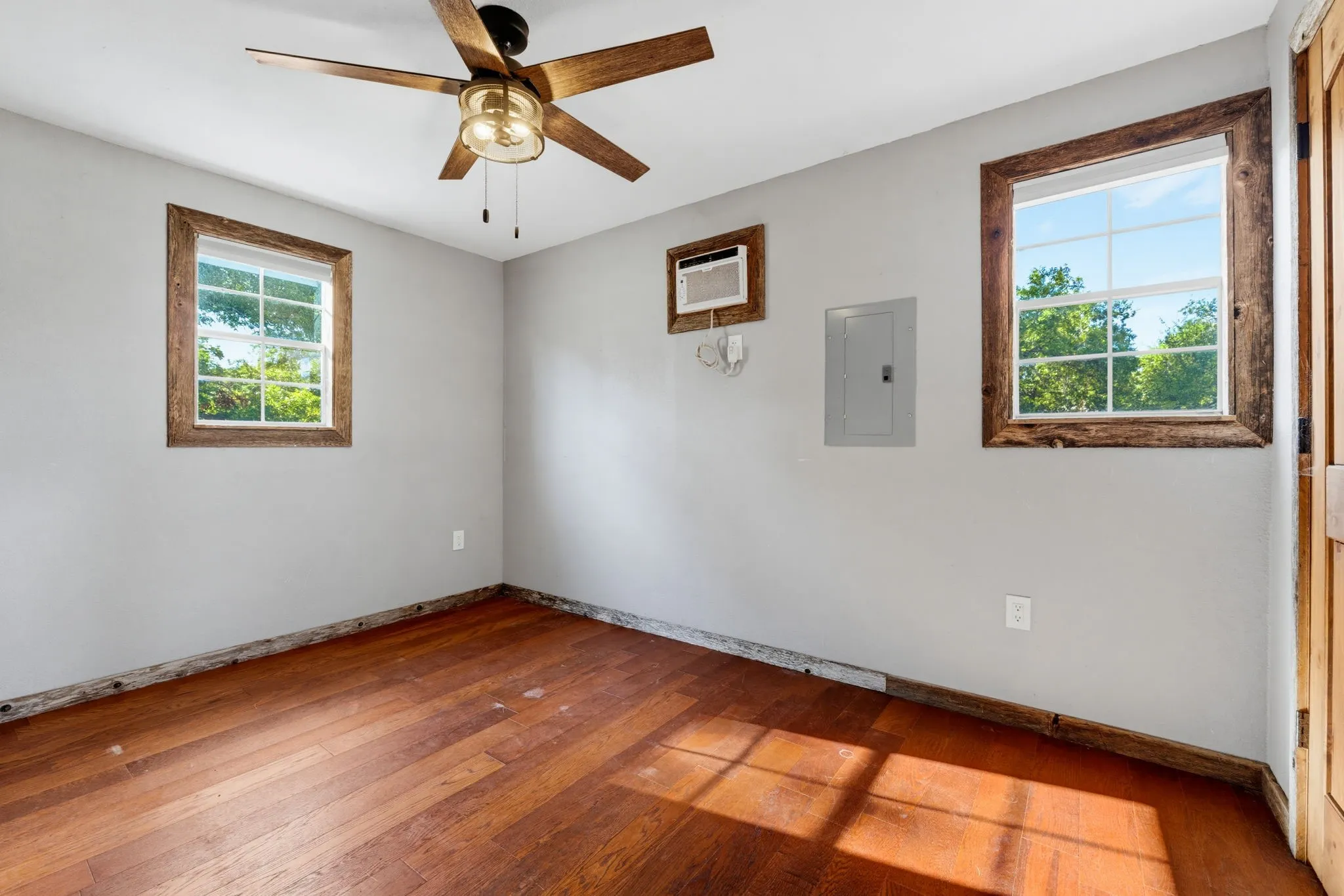 Wood-finish flooring, ceiling fan, two windows with wood-frame trim, and an electrical panel