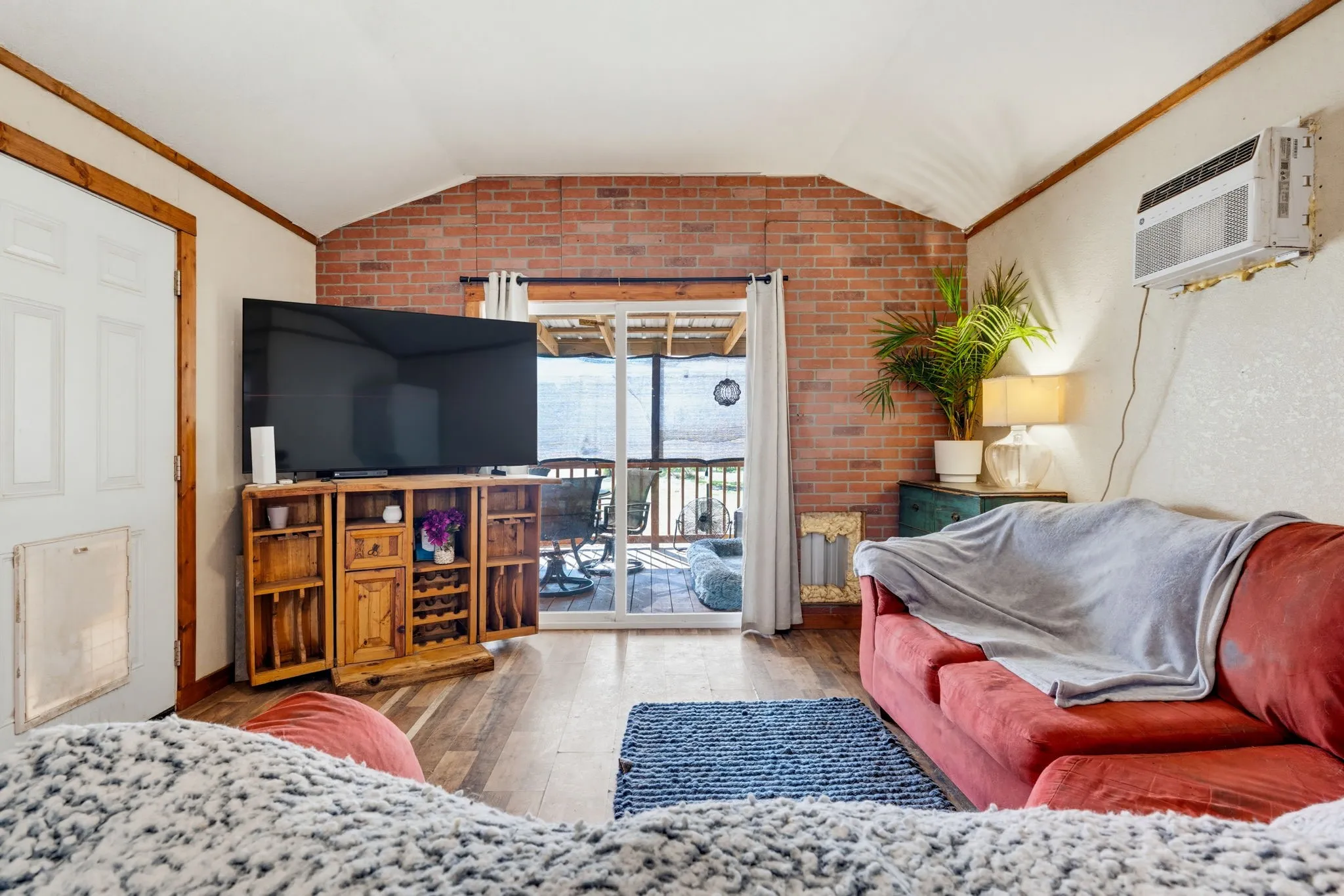 Vaulted ceiling with wood trim, brick accent wall, wood-finish flooring, and sliding glass door access to an outdoor area