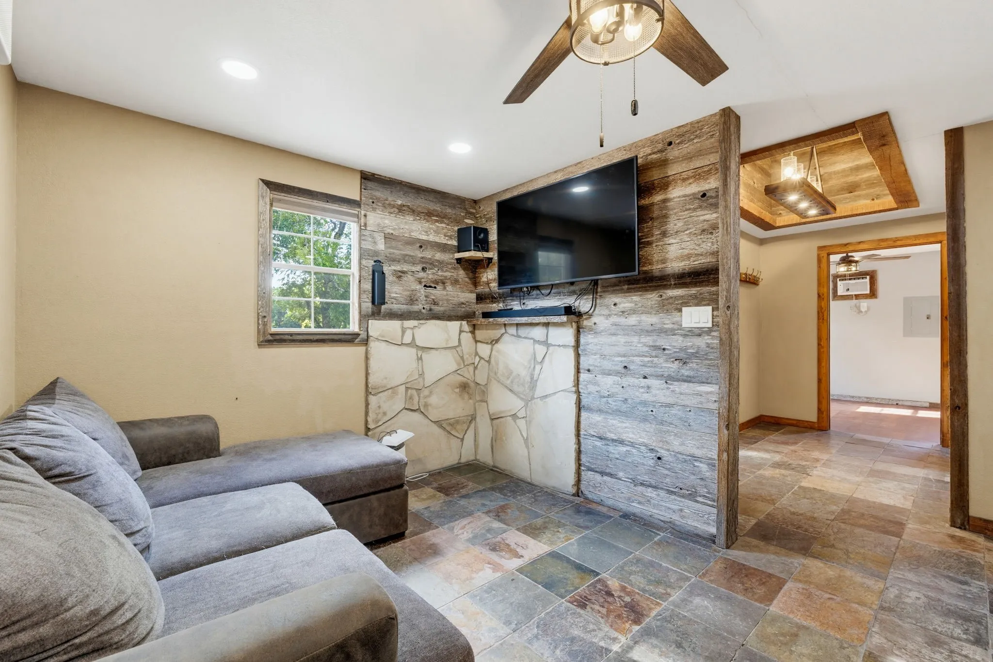 Recessed ceiling lighting, stone flooring, wood-paneled accent wall, wood-paneled ceiling fan, and exposed wood ceiling beams