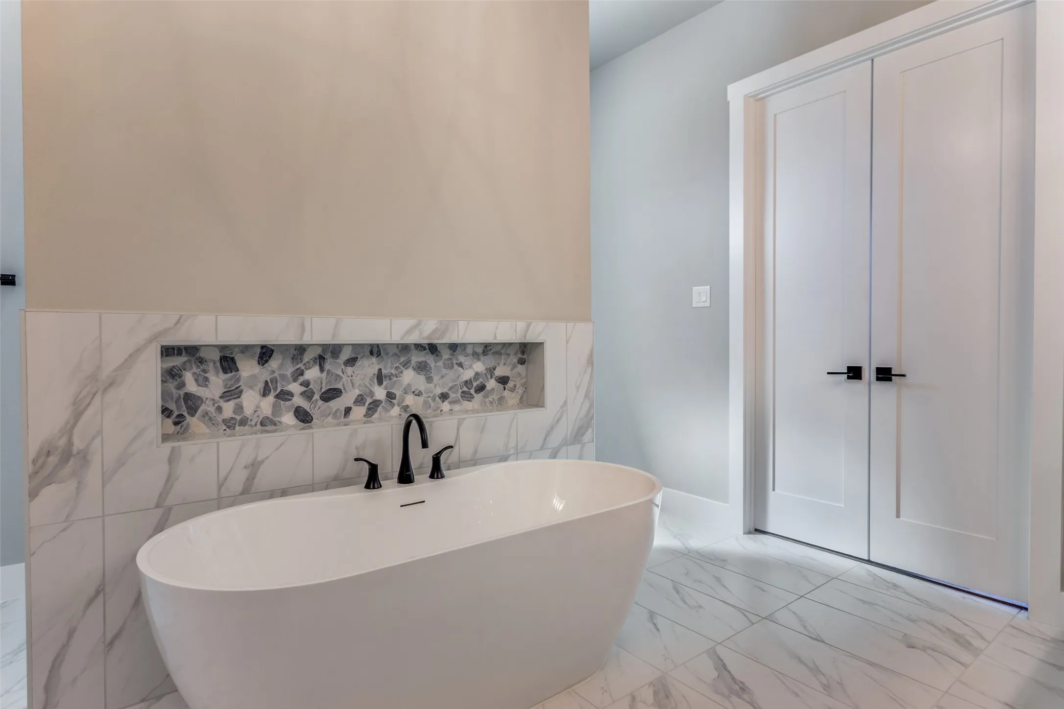 Freestanding soaking tub with matte black fixtures, marble-look tile surround, and a recessed mosaic shelf