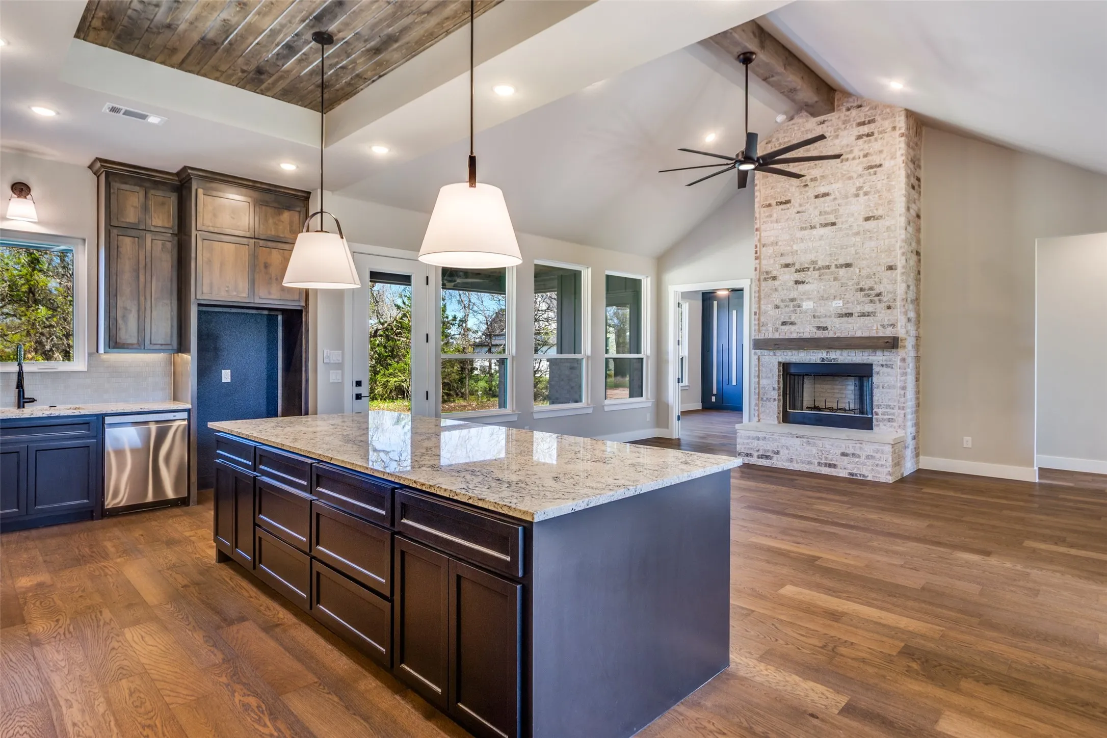 Vaulted ceilings with exposed beams, wood-finish flooring, a large kitchen island with a stone countertop, stainless steel appliances, and a brick fireplace