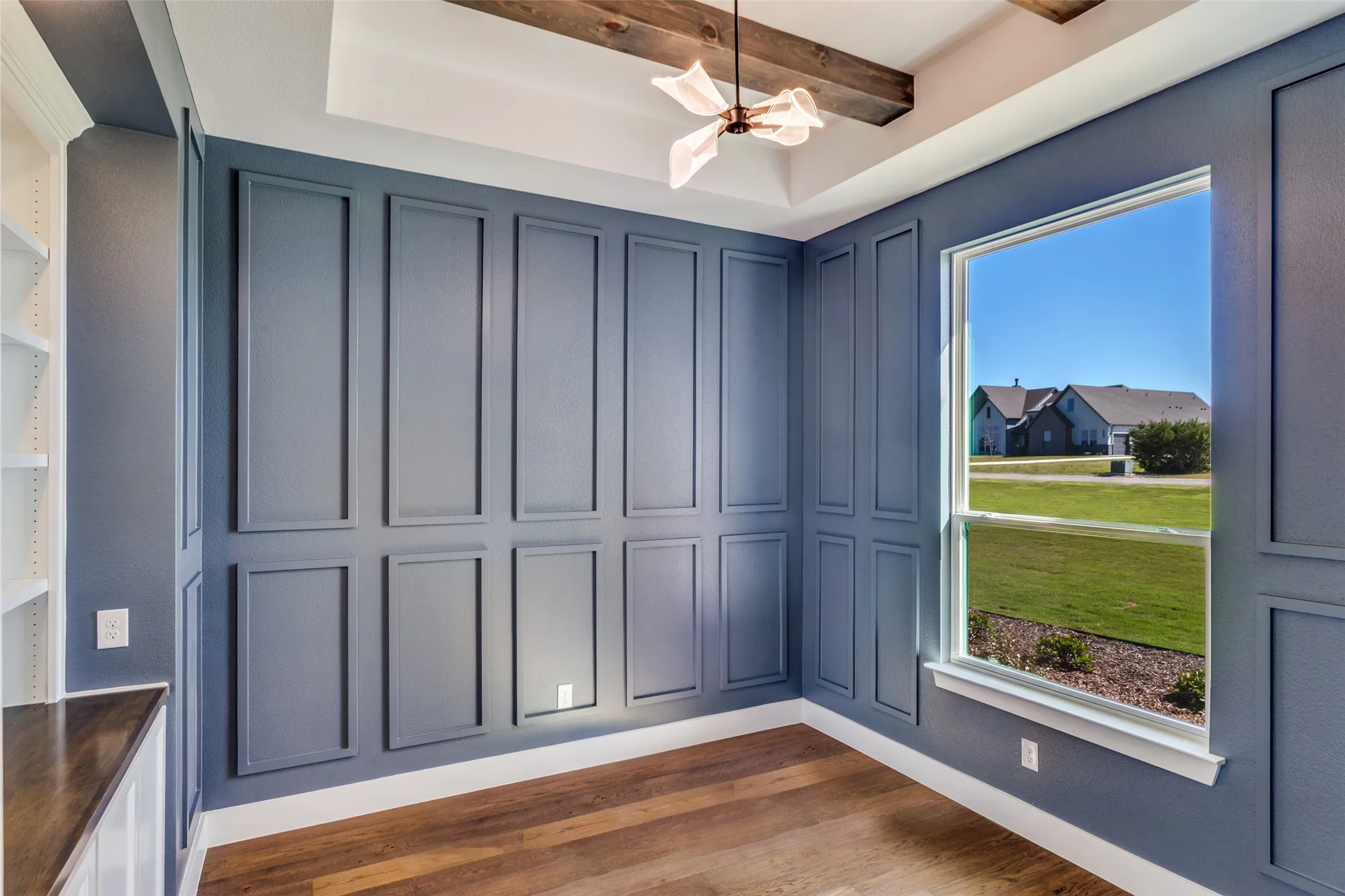 Tray ceiling with exposed wood beams, wood-finish flooring, built-in shelving, picture frame wall molding, and a single window
